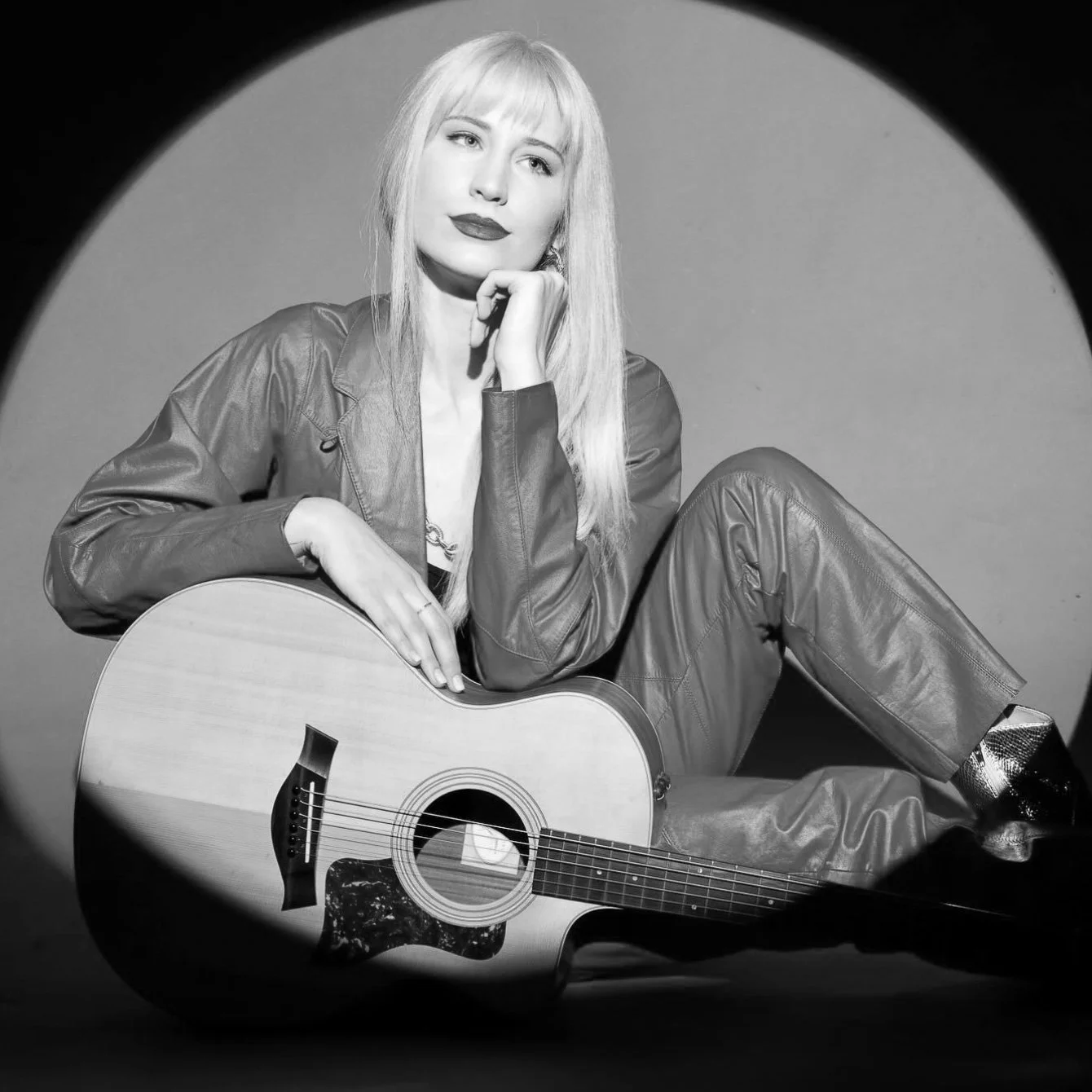 A black-and-white photo of a woman with long blonde hair, sitting with a guitar in her lap. She is wearing a leather jacket and matching pants, with her hand resting on her chin and gazing thoughtfully.
