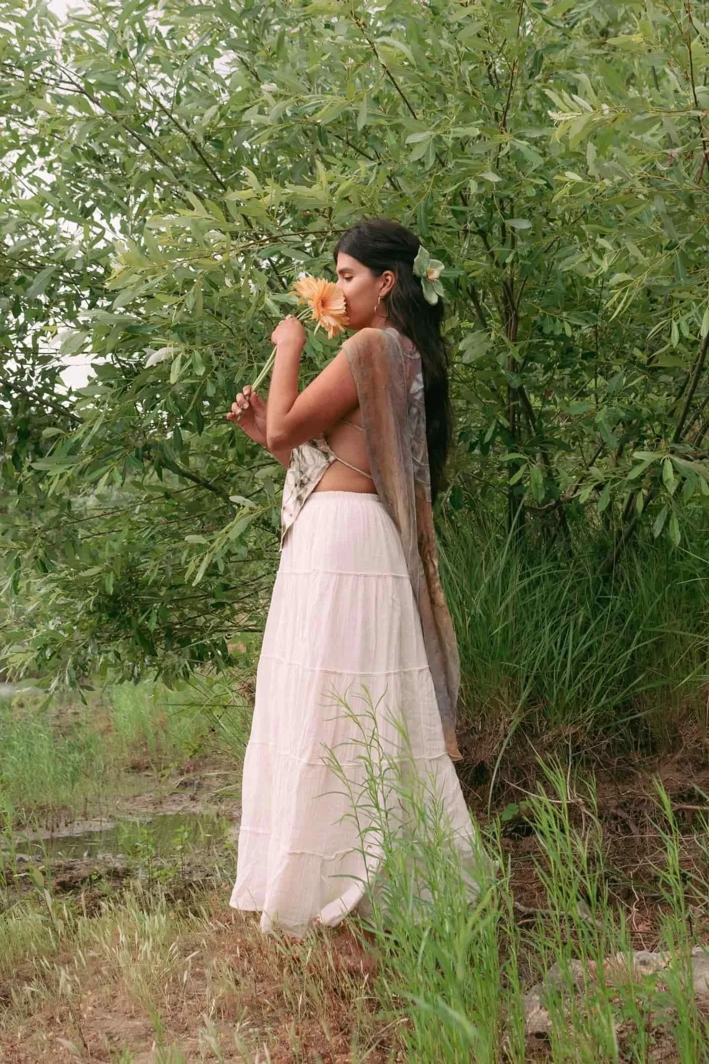 A woman with long dark hair, wearing a beige and white dress, is standing in a lush green outdoor setting, holding a peach-colored flower near her nose, with a large green flower tucked behind her ear.