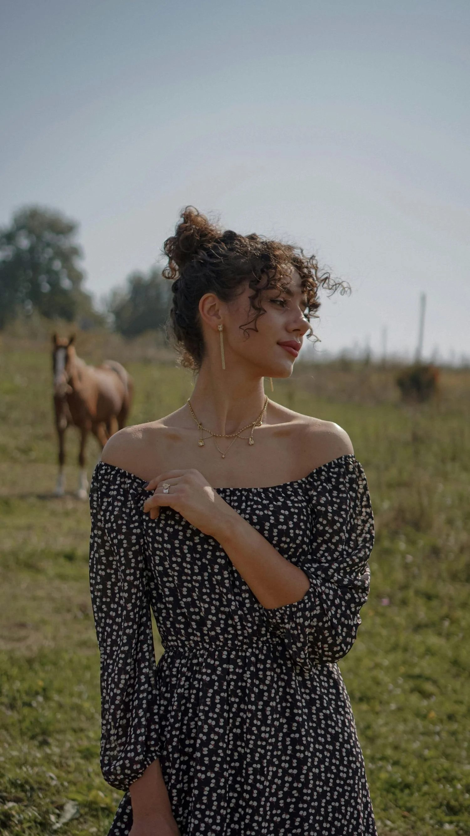 A woman with curly hair in a bun wearing an off-the-shoulder black and white dress and gold jewelry stands in a pasture with horses in the background.