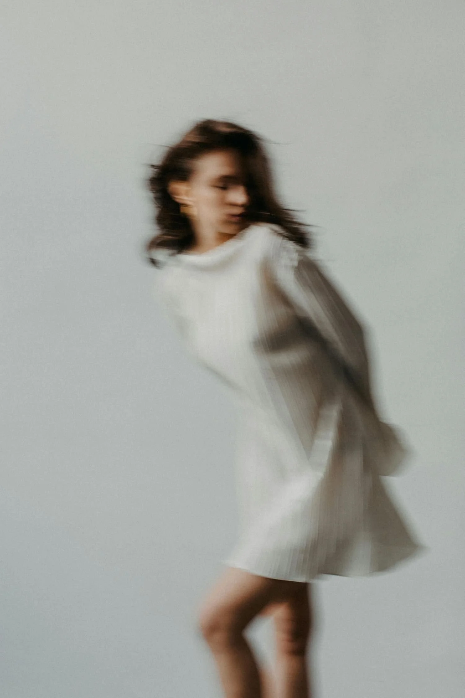 An artistic studio photo of a woman in a white dress leaning over with her hands behind her back and looking to the side. The photo has some motion blur