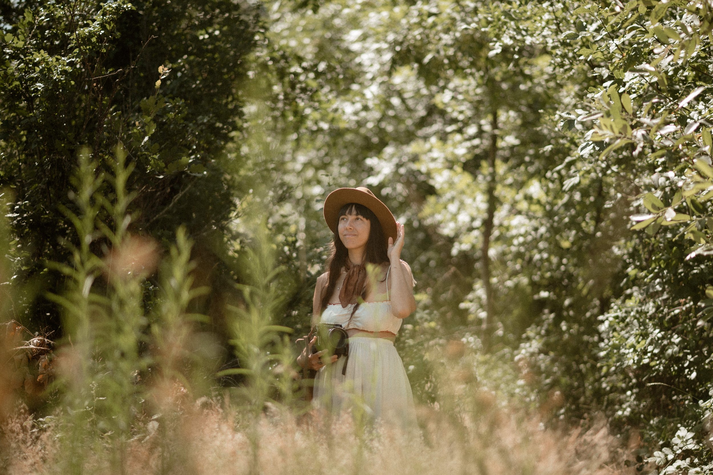 Woman in a hat standing in a lush, green outdoor setting with sunlight filtering through the trees.