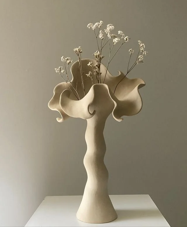 A beige, abstract ceramic vase with a wavy, organ-like design, holding small white dried flowers, displayed on a white surface against a plain neutral background.