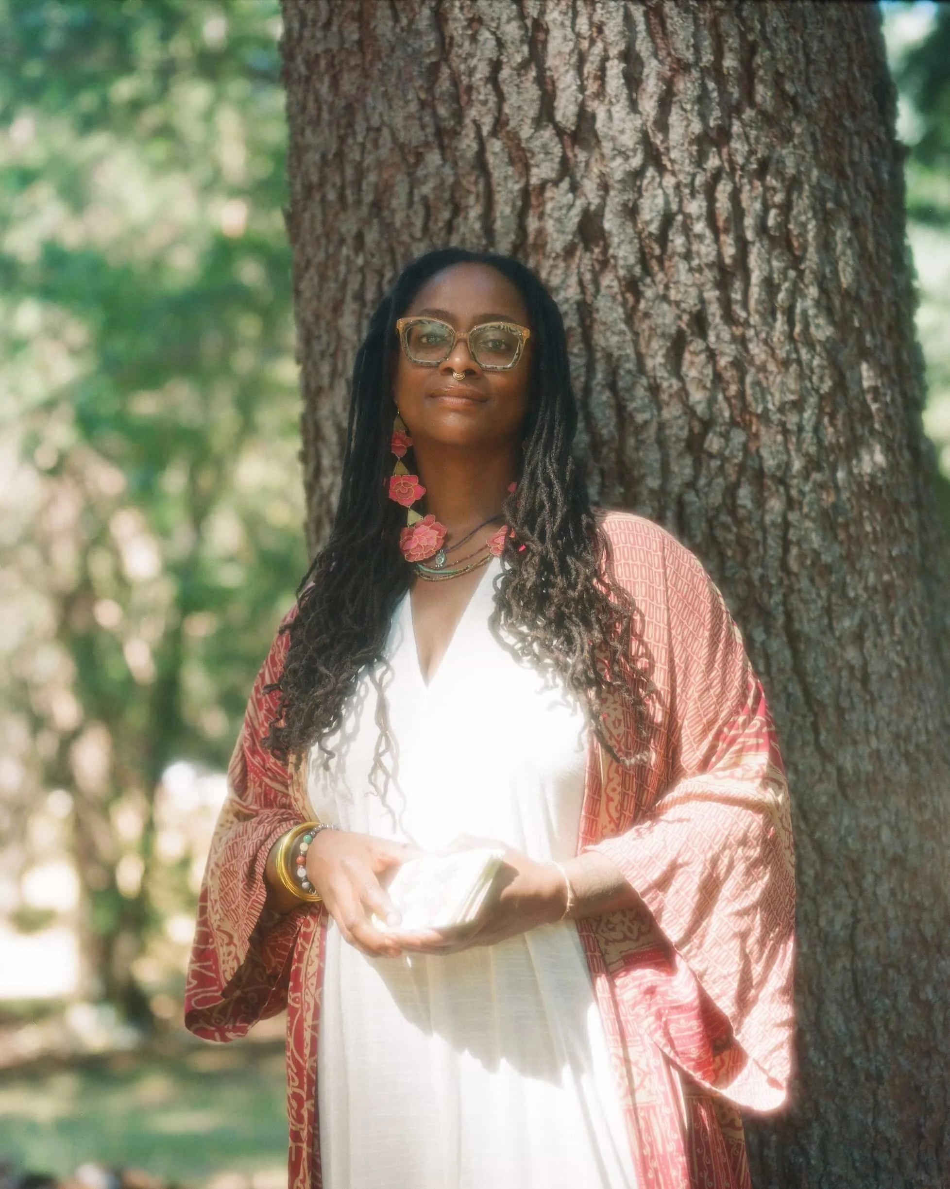 A woman with dark, dreadlocked hair and glasses leaning against a tree outdoors during daytime. She is wearing a light-colored dress and a patterned, pinkish-red shawl, with colorful jewelry on her wrist and neck. She is holding a book, and the background is blurred with green foliage.