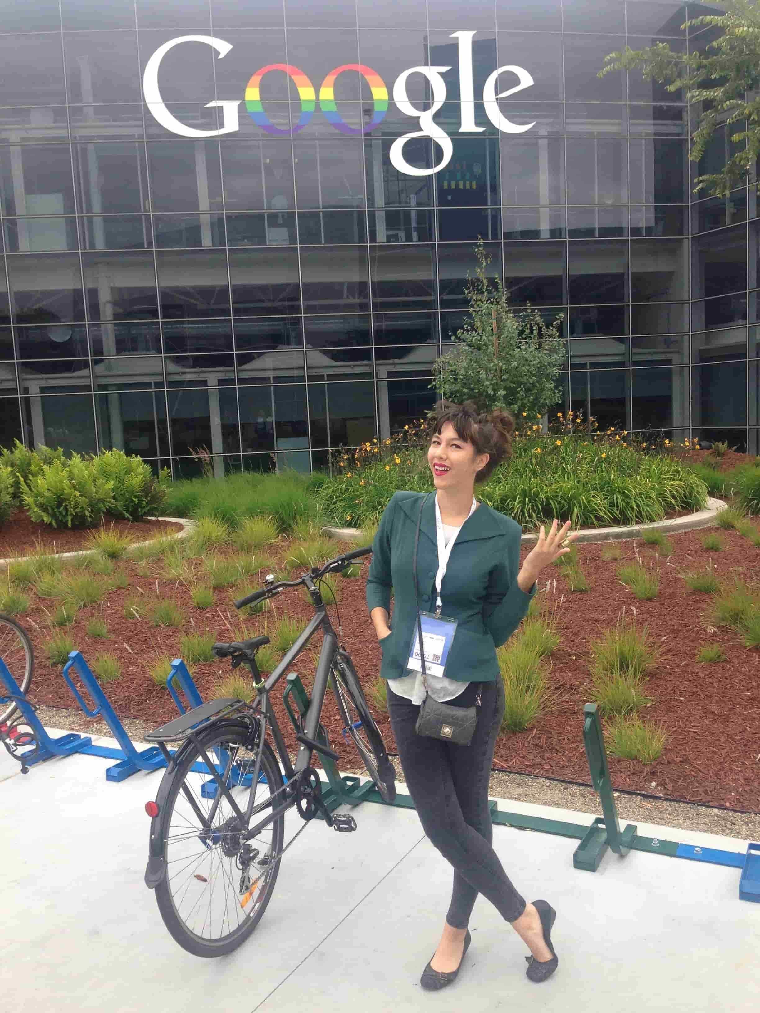 A woman with curly hair, dressed in dark pants and a green blazer, standing outdoors next to a bicycle rack, smiling and waving, in front of a modern building with Google logo and a garden with small plants.