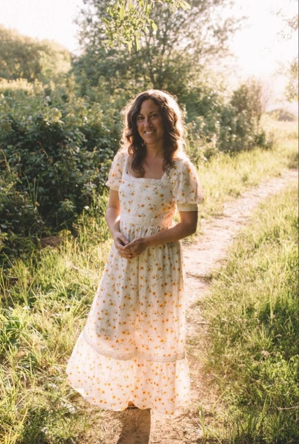 A woman standing outdoors on a dirt path beside lush greenery, wearing a long, floral dress with short sleeves, smiling, illuminated by warm sunlight.