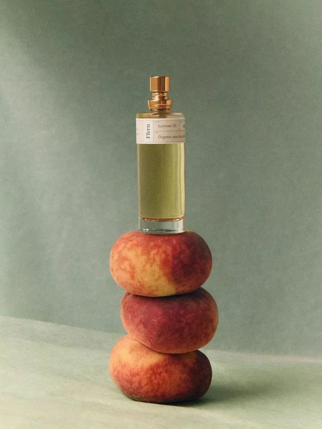 Stack of four peaches with a perfume bottle on top, set against a neutral background.
