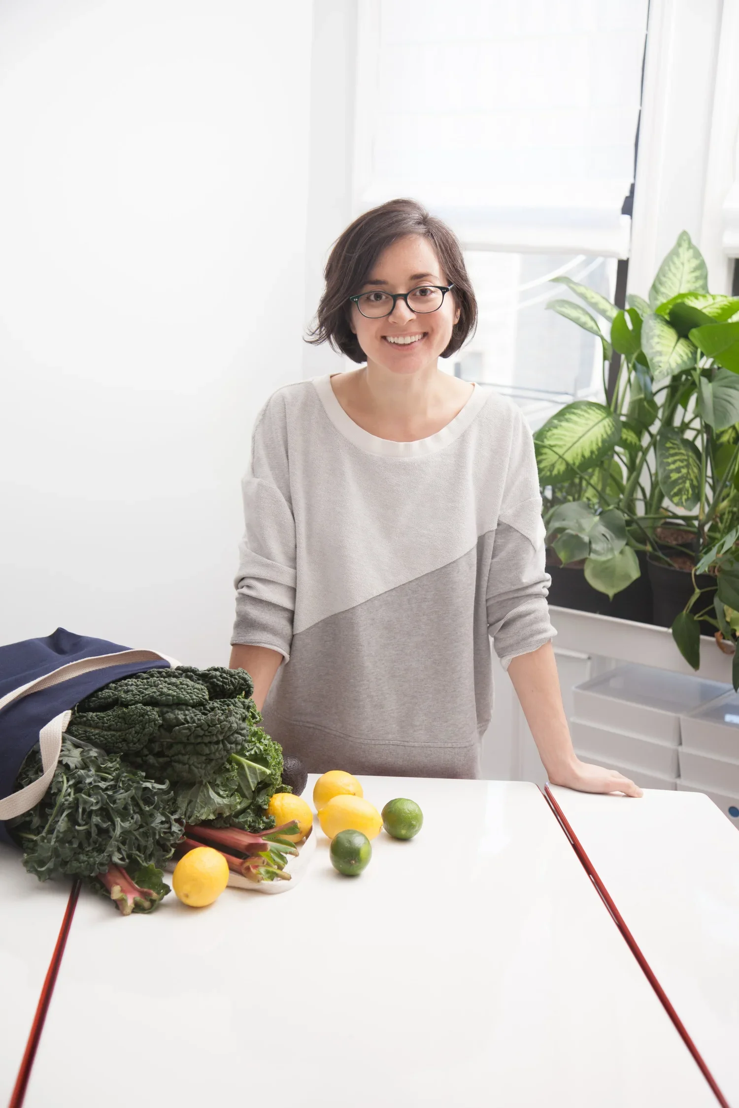 A woman with glasses and short brown hair smiling at a table with fresh vegetables and citrus fruits next to her, in a bright room with a window and a green plant in the background.