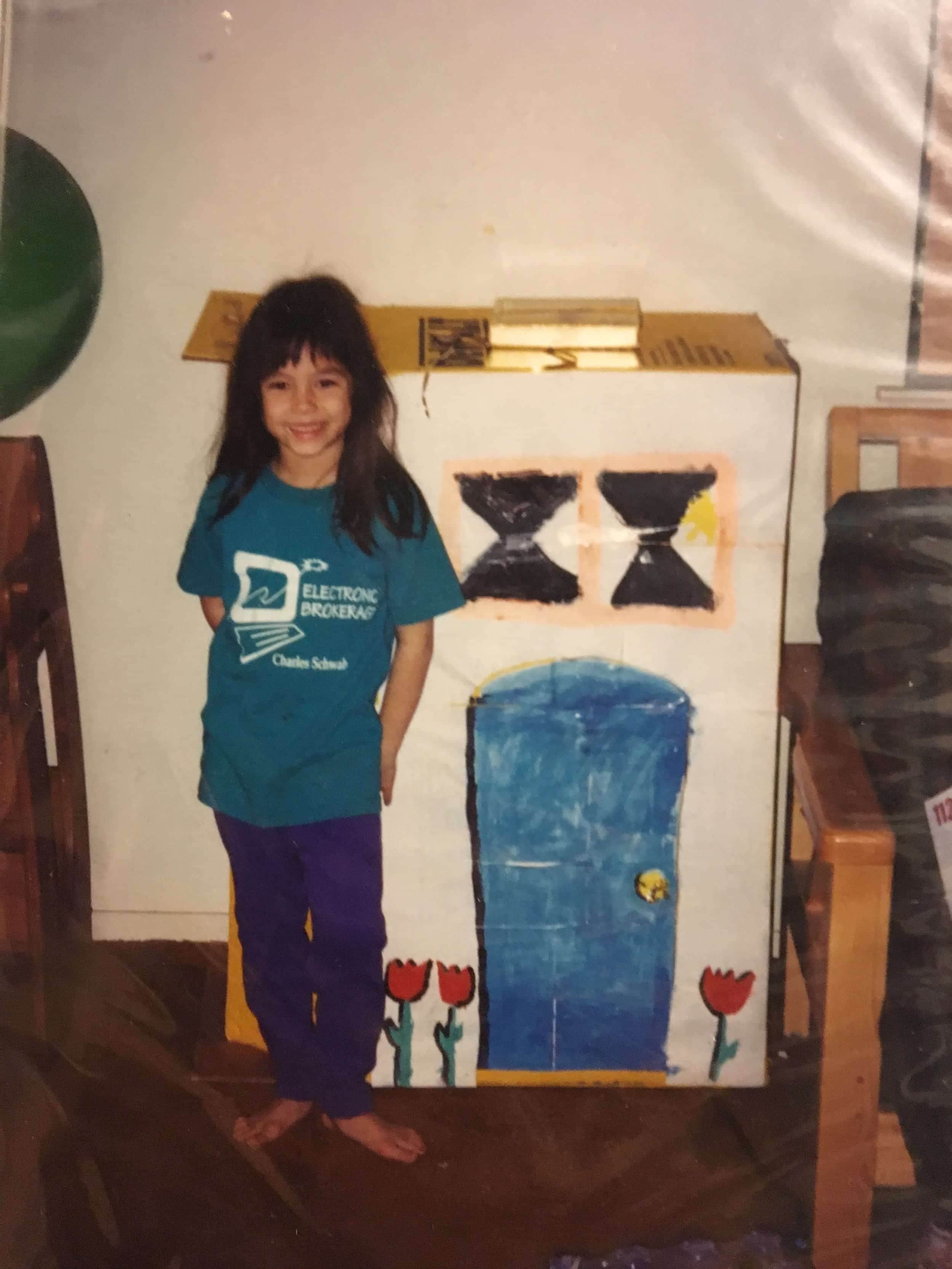 A smiling girl standing next to a painted wooden stand decorated with tulips and cartoon character designs, in a room with wooden furniture and a white wall.