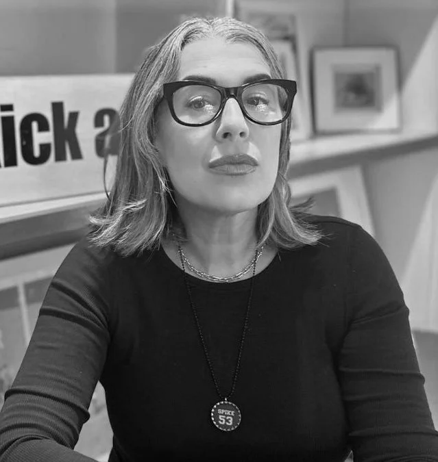 A woman with shoulder-length hair wearing glasses, a black top, and a necklace with a pendant that reads 'SPIKE 53' sitting at a desk with framed pictures behind her.