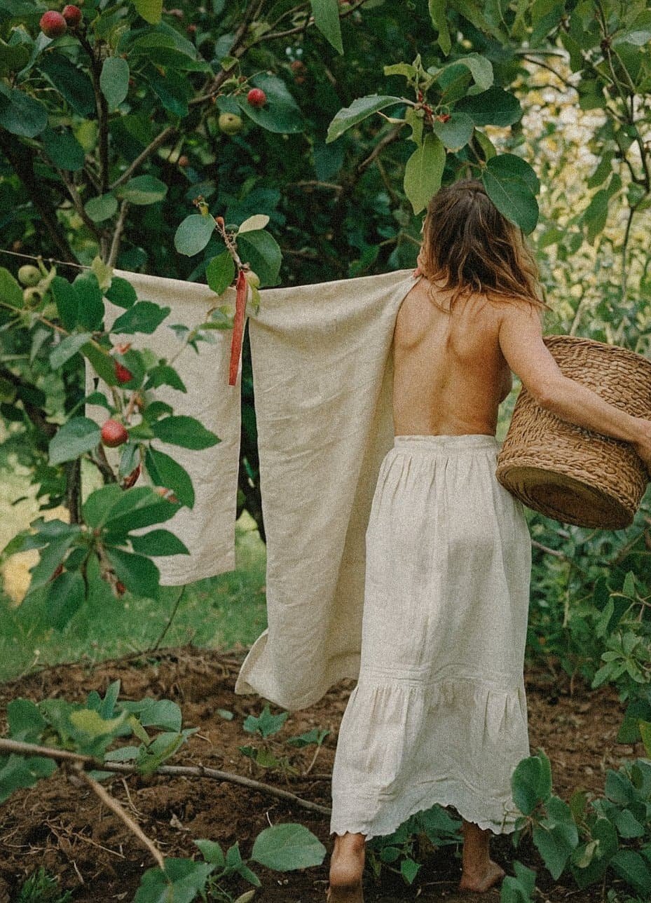 A woman with long hair, topless, wearing a long light-colored skirt, walking through a lush garden or orchard, holding a wicker basket, with a large cloth or sheet draped over her shoulder. The area is filled with green leafy trees and small reddish fruit.