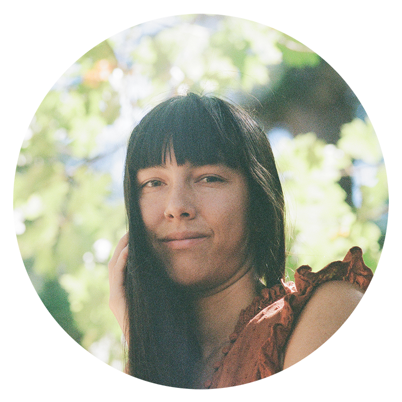 Young woman with long dark hair and straight bangs outdoors surrounded by green foliage, wearing a reddish-brown ruffled top, looking at the camera with a slight smile.