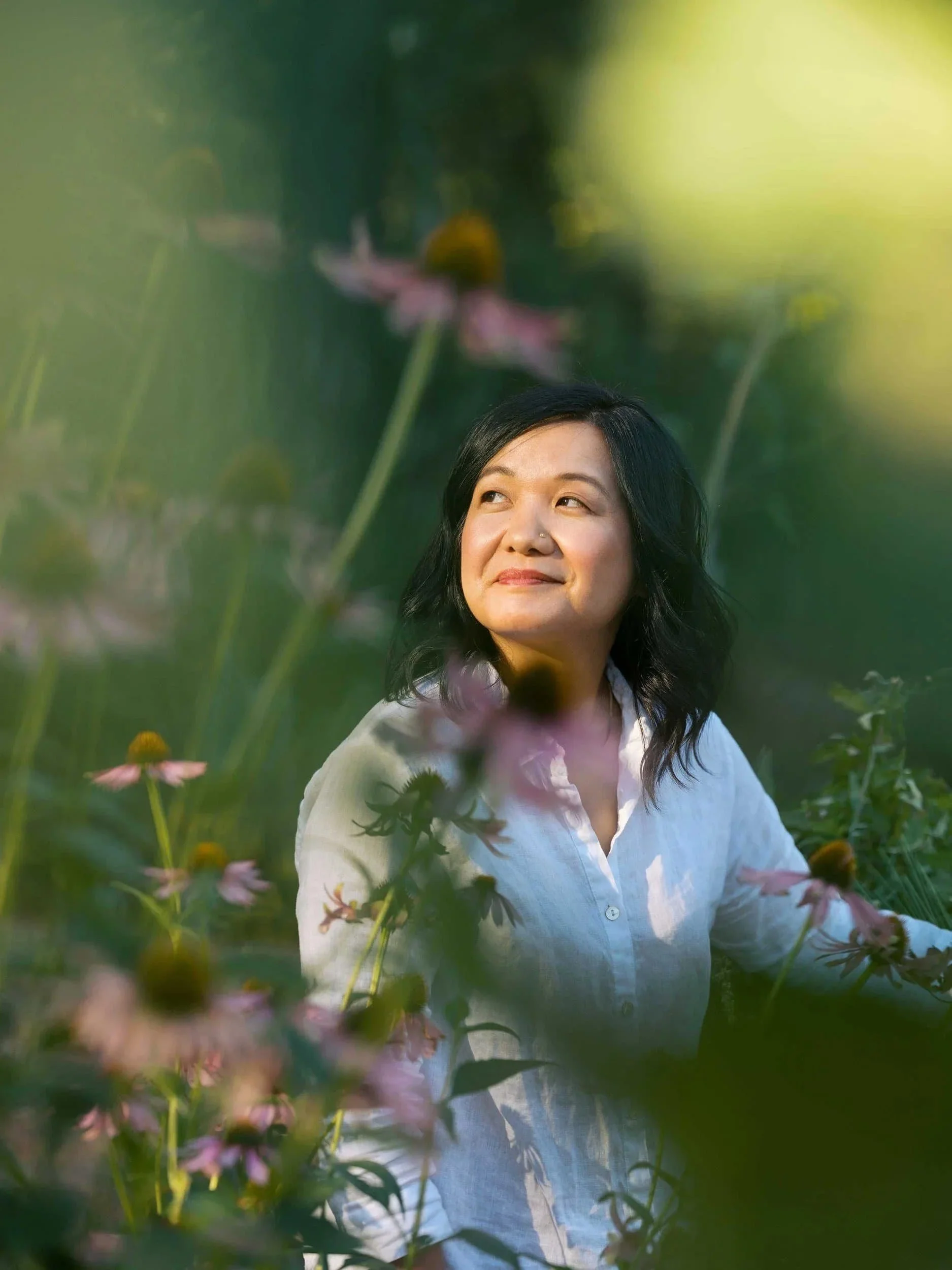 Female founder Danielle Tsi with black hair wearing a white shirt outdoors among green plants and pink flowers, looking upward with a smiling expression.