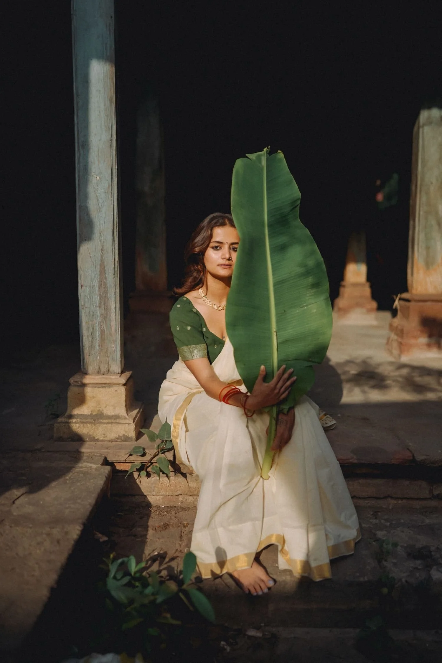 Woman sitting on steps holding a large green leaf, wearing traditional Indian attire, outdoors at night.