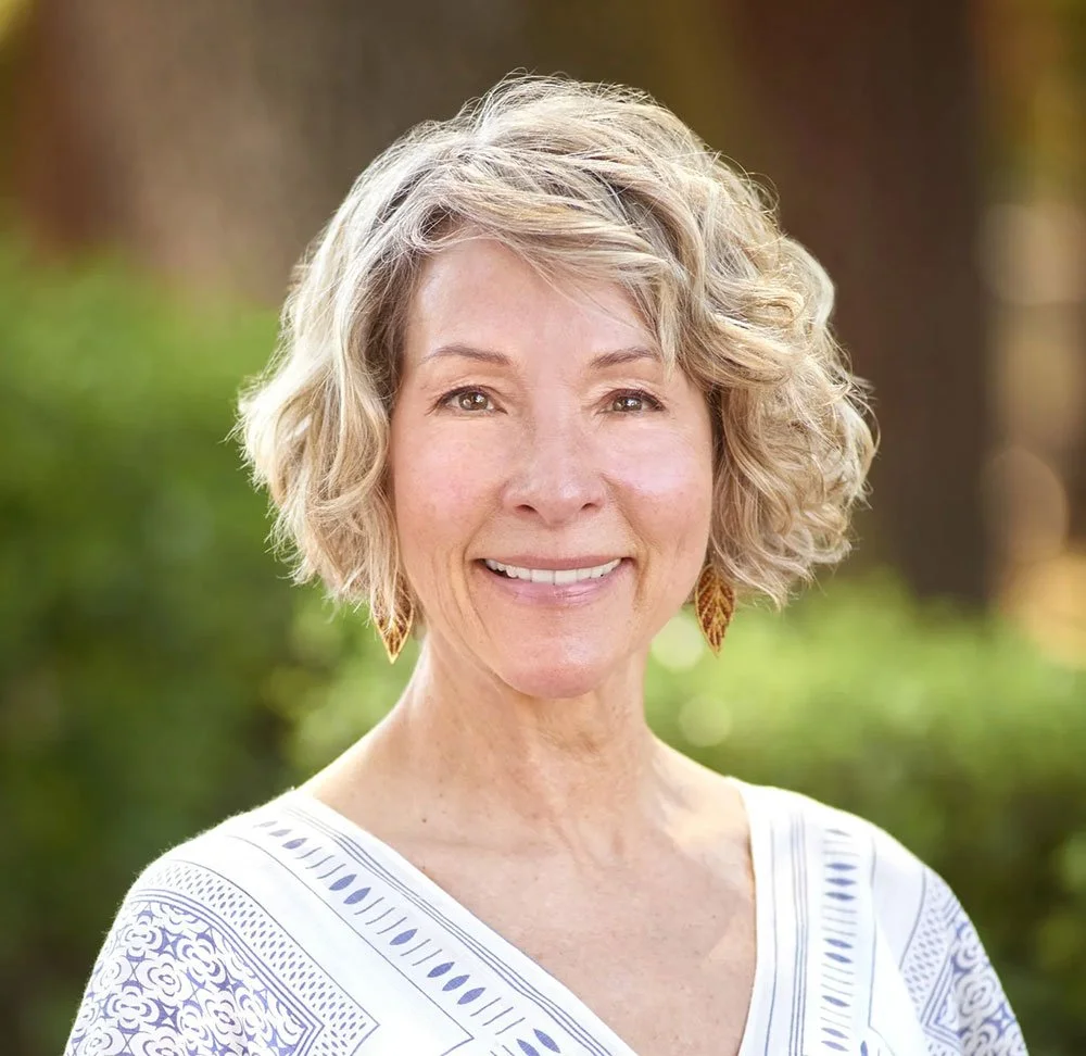 A middle-aged woman with short, curly blonde hair outdoors, smiling, wearing a white top with patterned embroidery and leaf-shaped earrings.