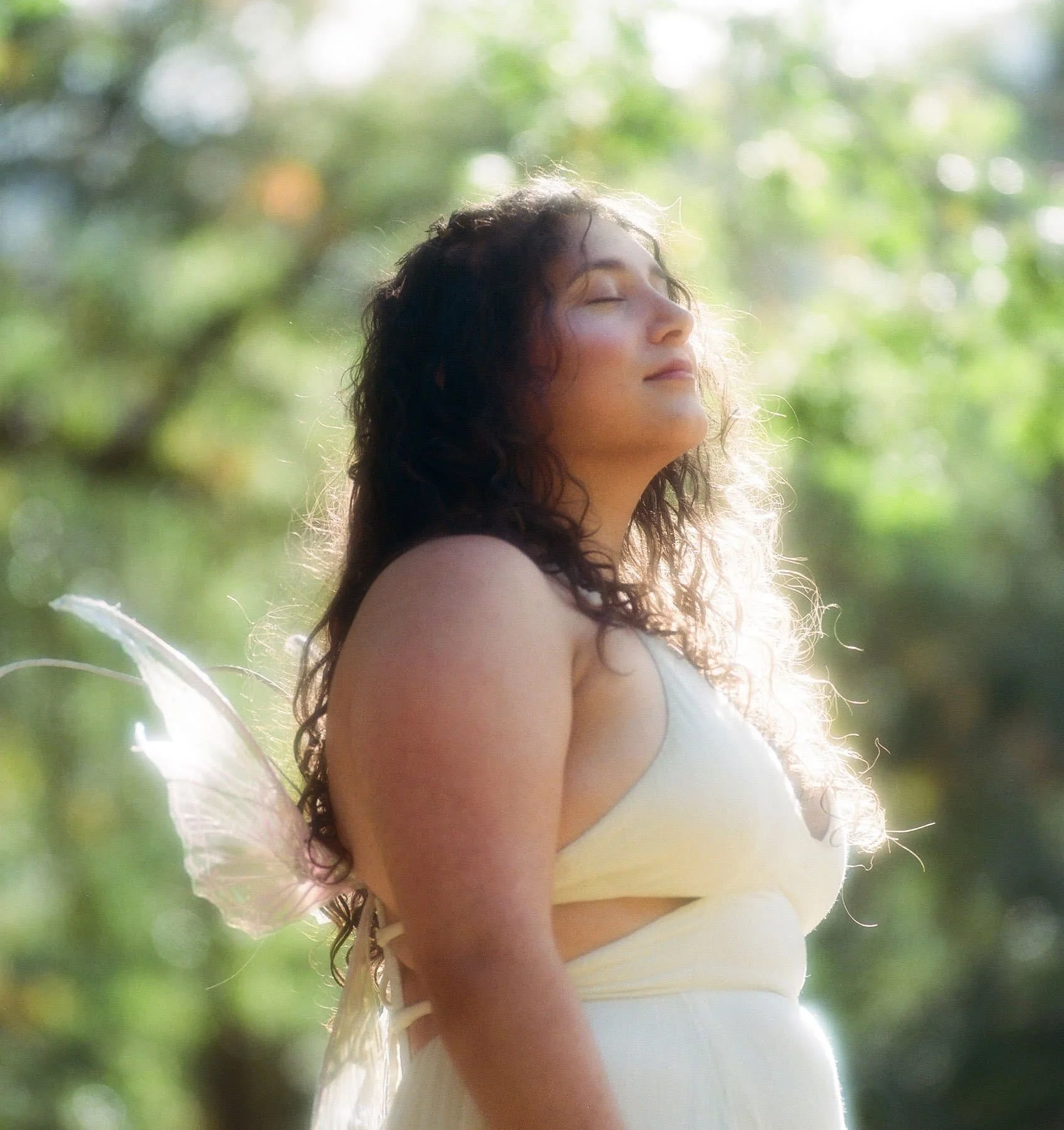 A woman with dark, curly hair and fair skin, wearing a yellow dress, outdoors with sunlight and greenery in the background, with her eyes closed and a peaceful expression.