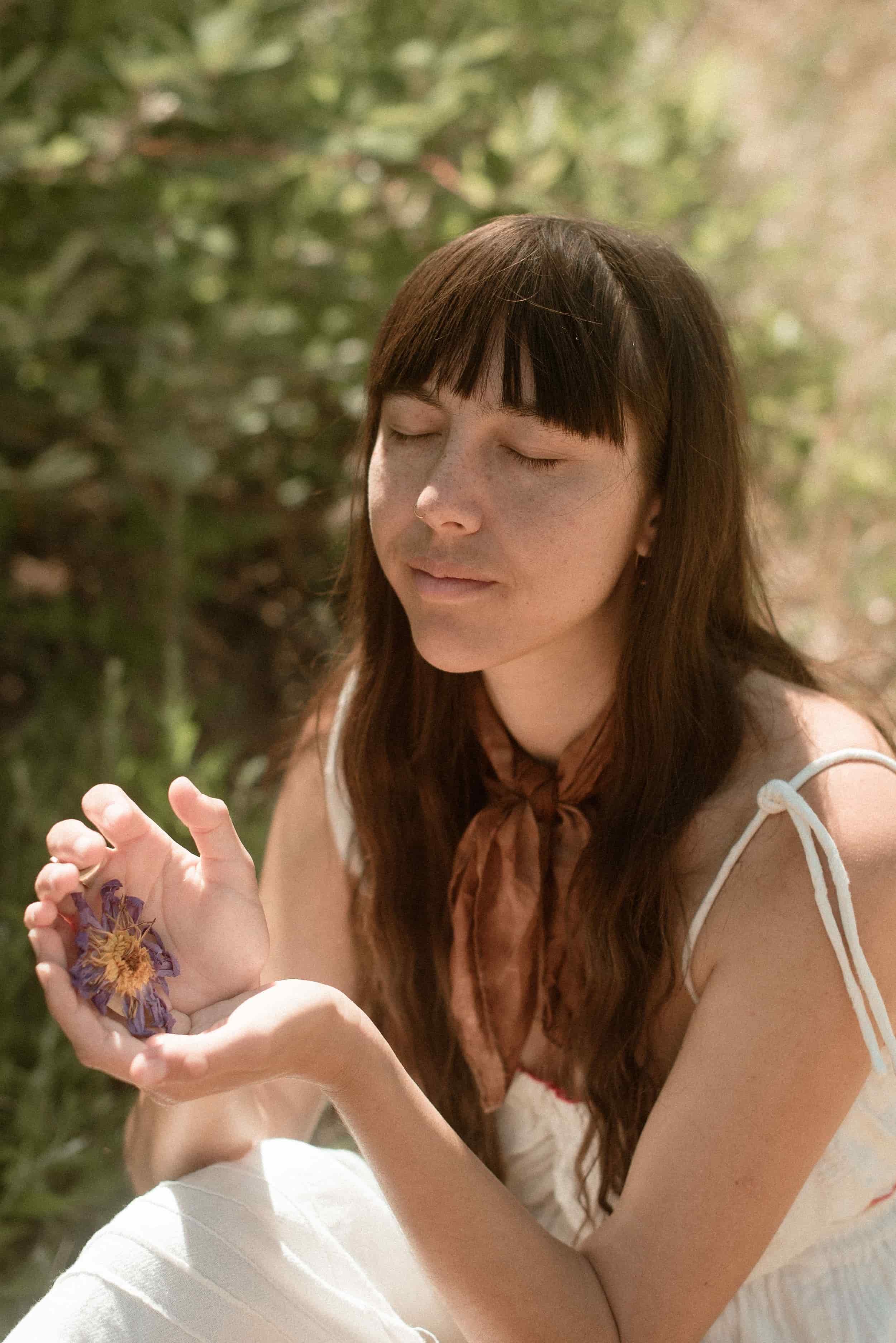 A young woman with long brown hair and freckles sitting outdoors with eyes closed, holding dried flowers in her hands, against a blurred green background.