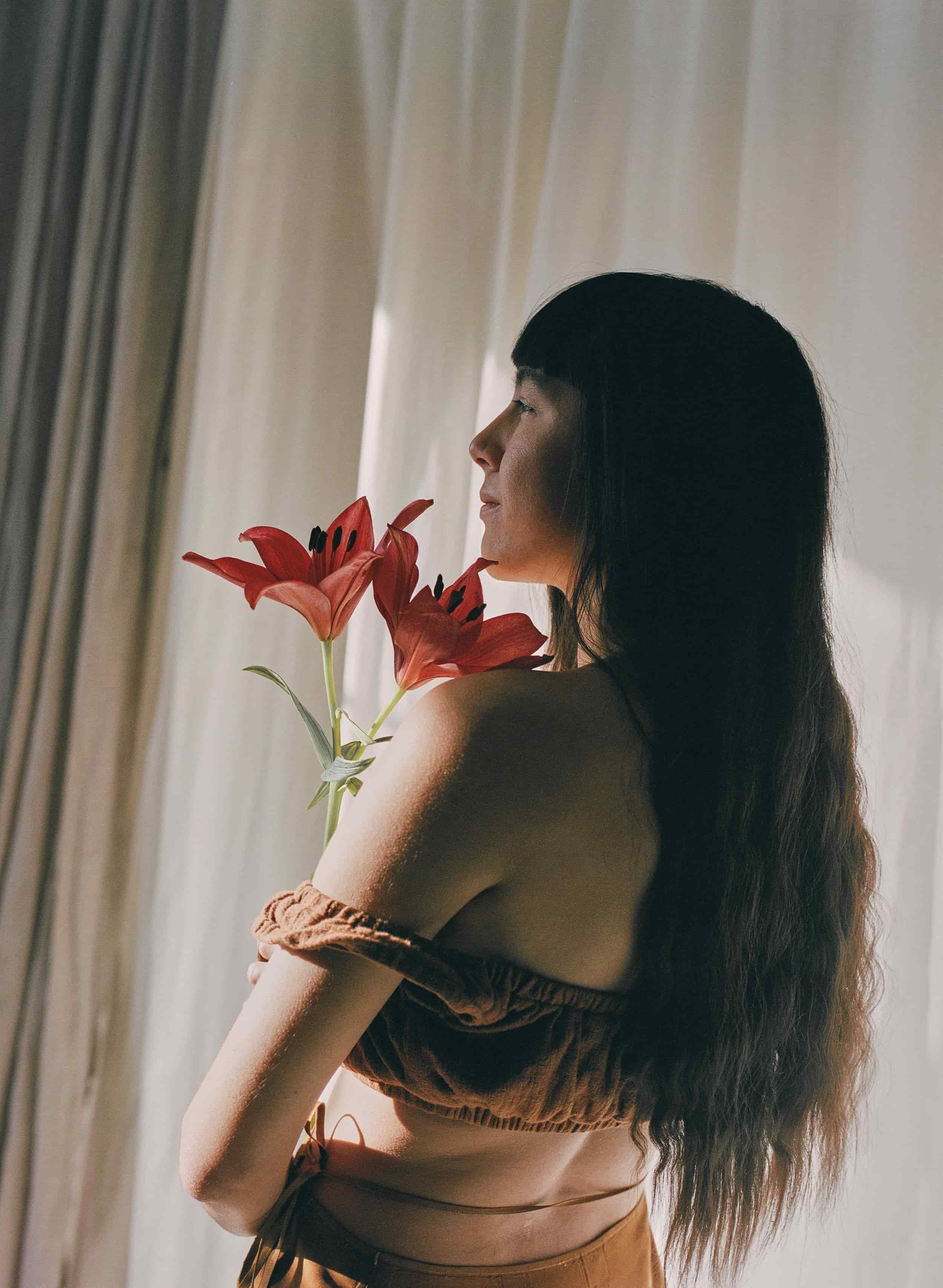 A woman with long dark hair looking to the side, holding a bouquet of red lilies near her face, standing in front of a sheer curtain with soft natural light.