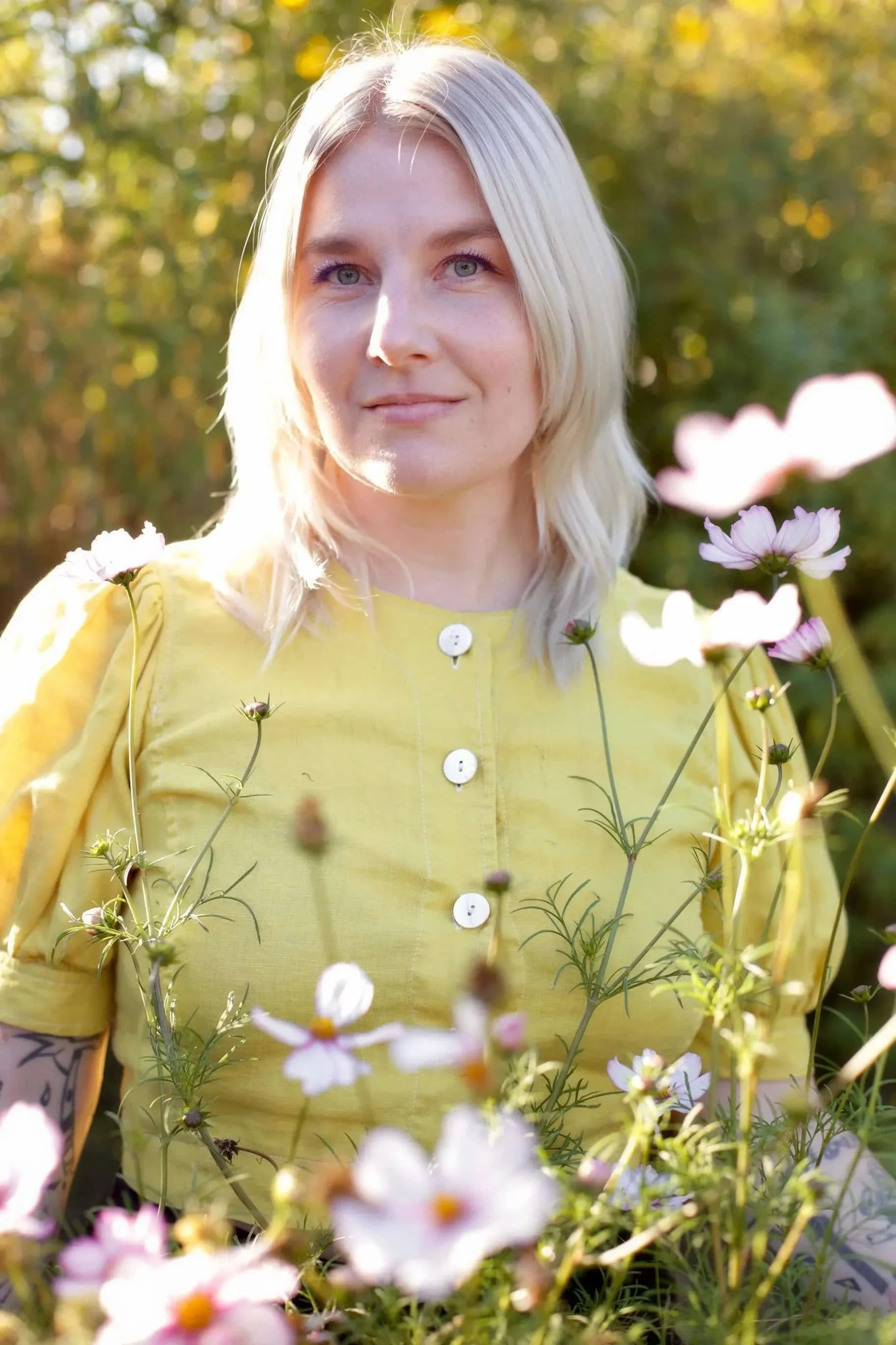 A woman with blonde hair and a yellow dress stands outdoors among pink flowers with a blurred green background.