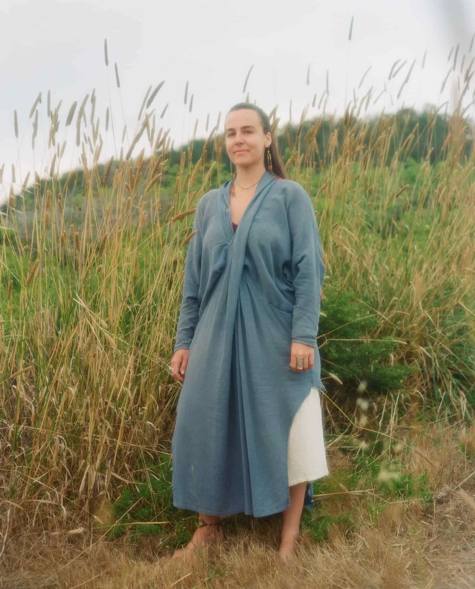 Female founder Emma Marie standing barefoot in tall grass and wildflowers with a slight smile, dressed in a blue shawl over a dark top, with earrings and necklaces, in a natural outdoor setting with a cloudy sky.