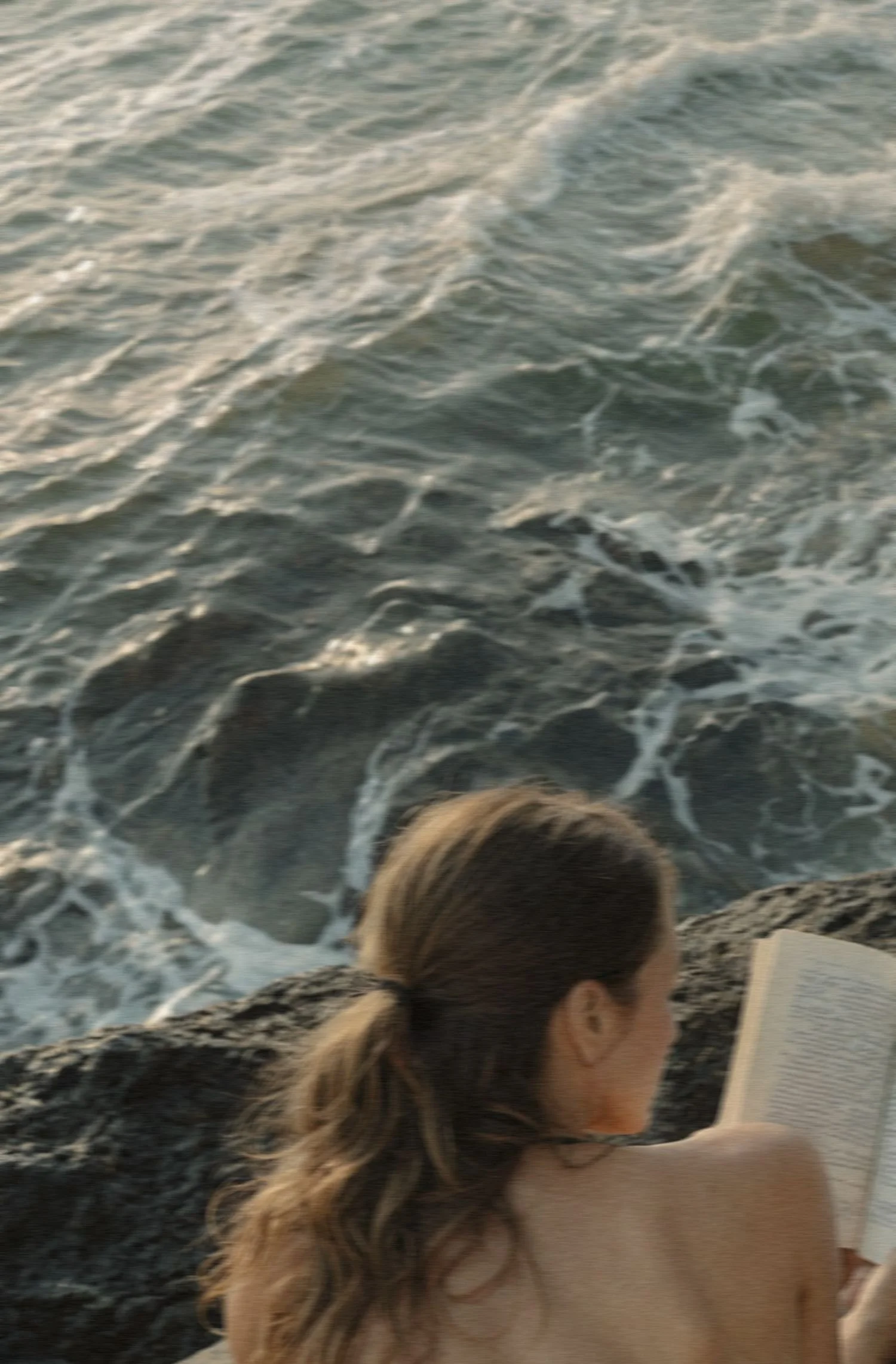 Woman with blonde hair reading a book on the beach. Waves in the ocean in the background.