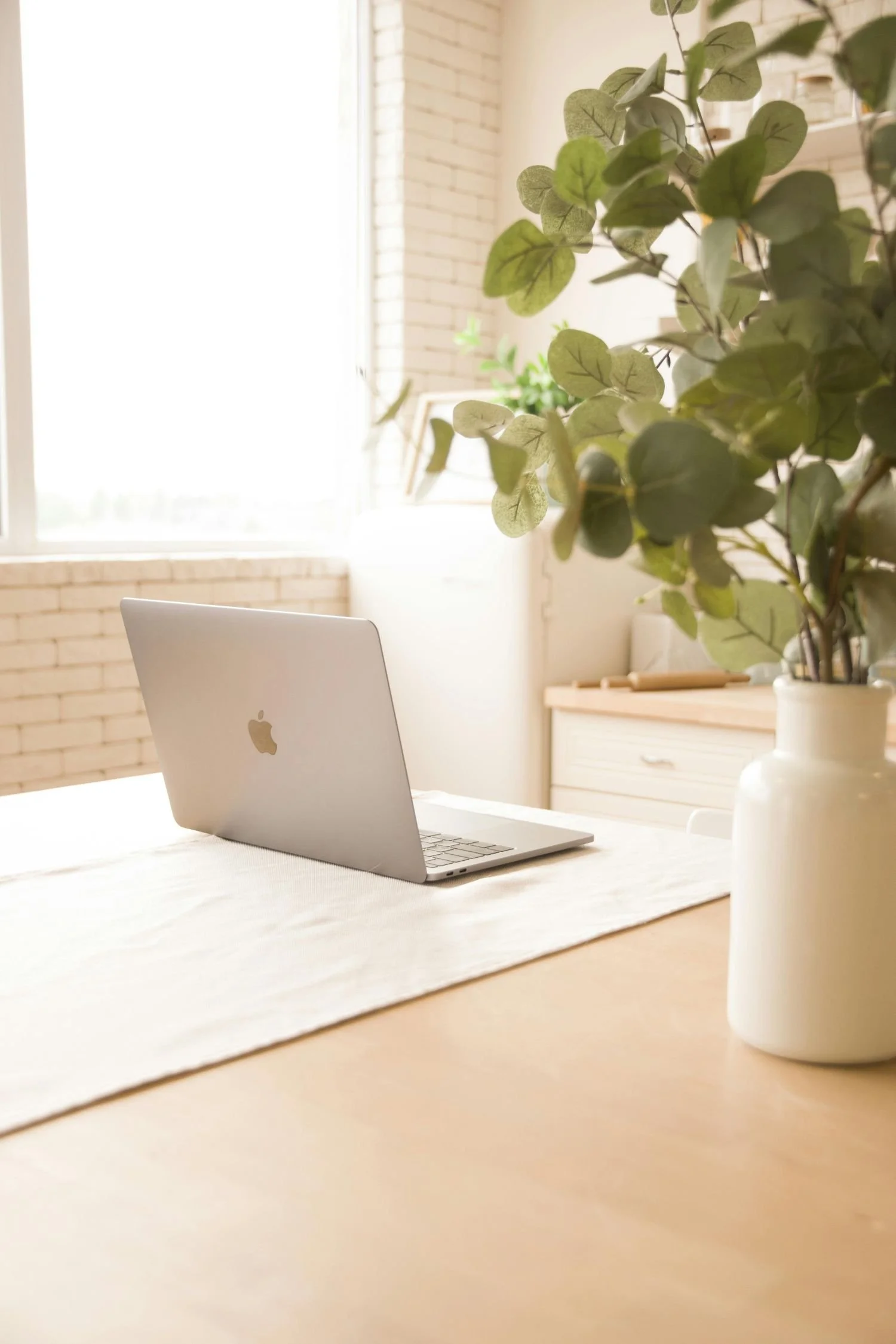 A silver MacBook laptop on a table with a white tablecloth, a white vase with green foliage, and a wooden dresser in a bright room with large windows.