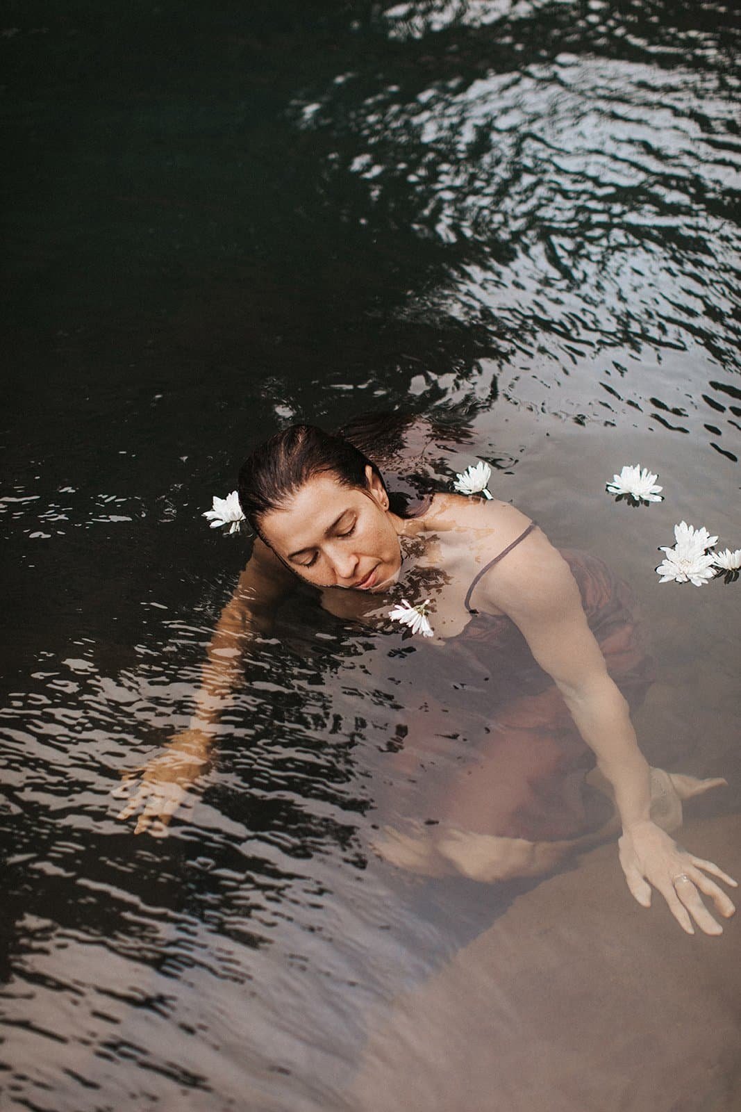 Female founder Sasha Gocloski with dark hair relaxing in water, surrounded by floating white daisies, with her eyes closed and a peaceful expression.