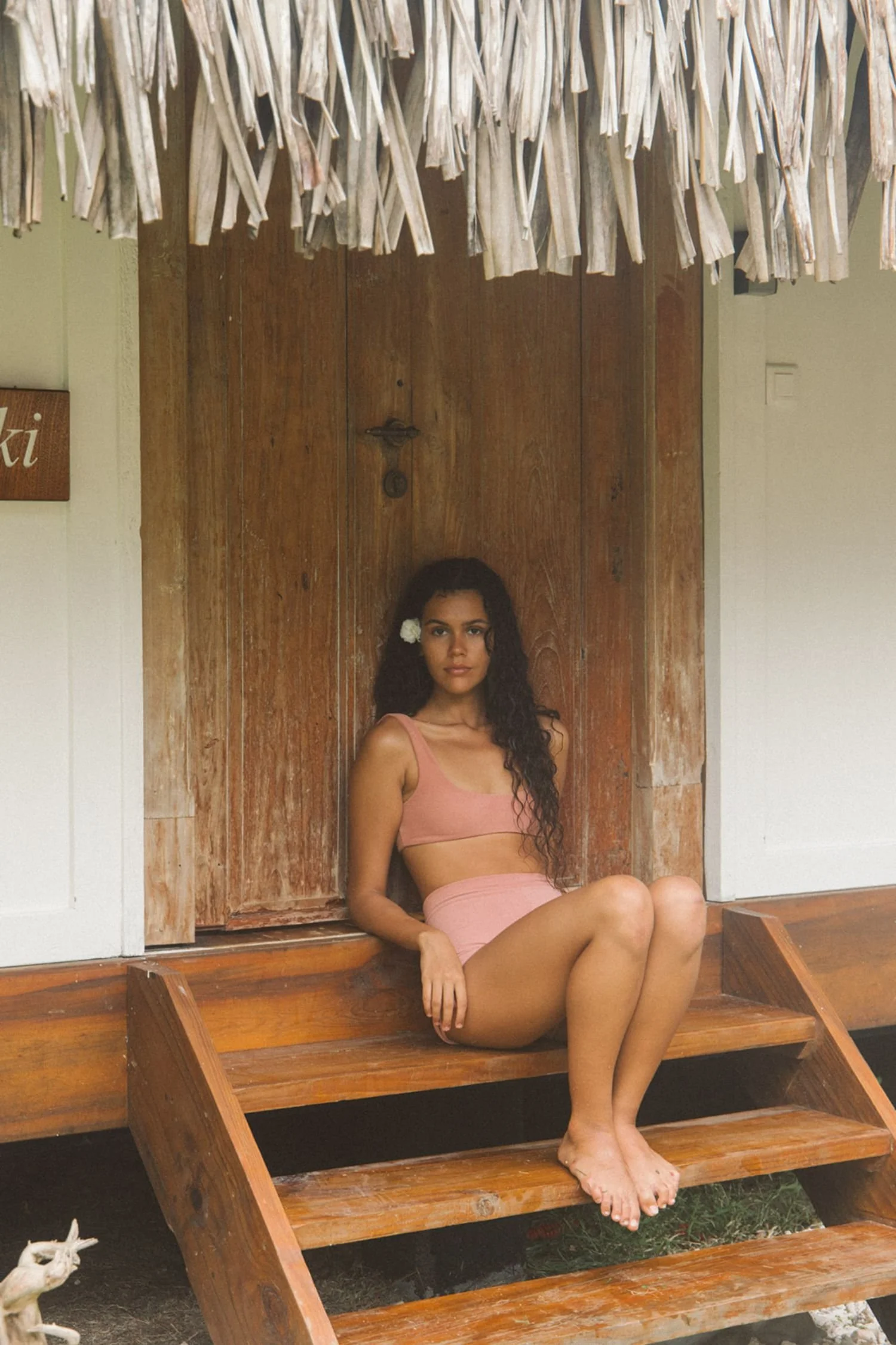 A woman with long curly hair and a flower in her hair is sitting on a wooden bench in front of a wooden door, wearing a pink sports bra and matching pink shorts. She is barefoot with her legs crossed, and a goat is visible in the bottom left corner of the image.