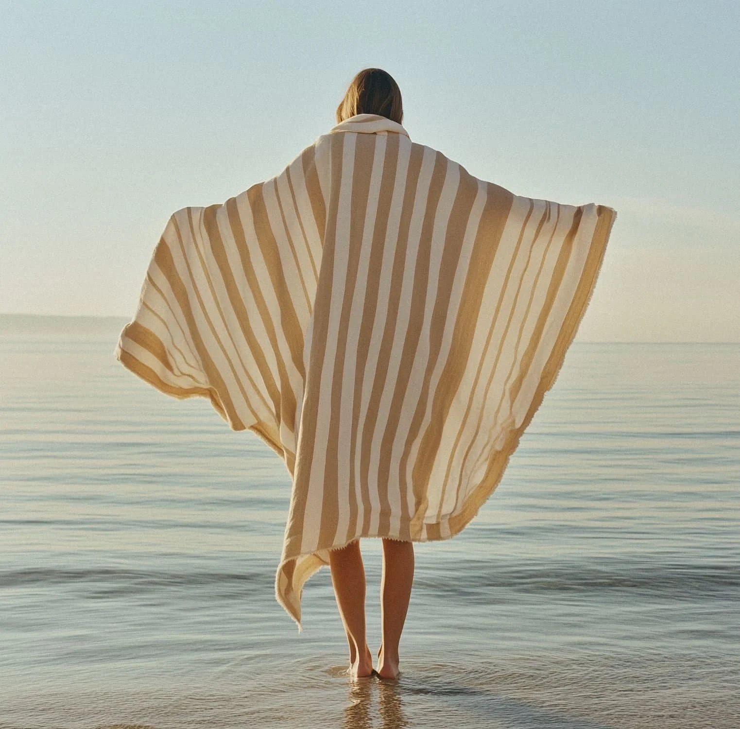 A person standing in shallow water at the beach, facing the ocean, wrapped in a large striped towel or blanket with outstretched arms.