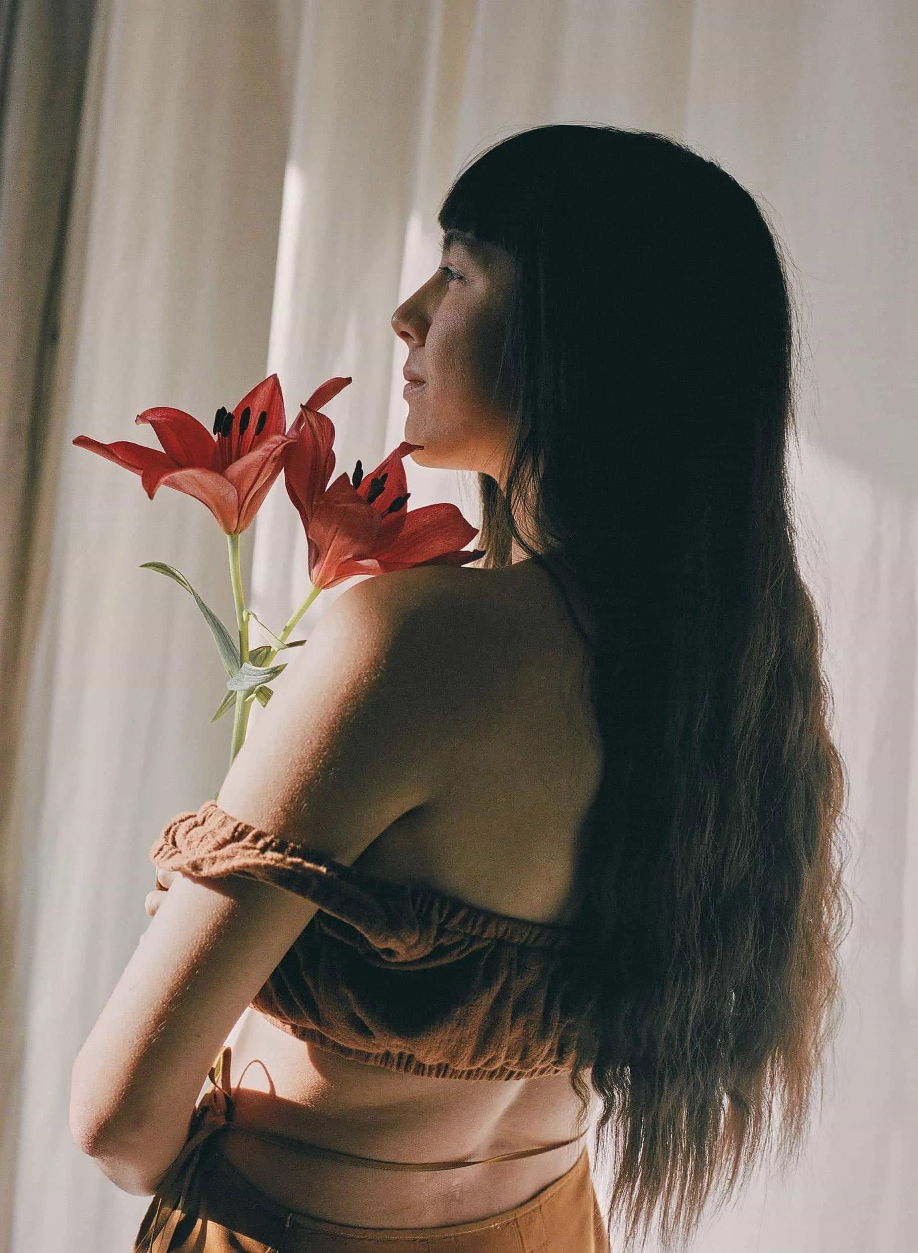 A woman with long dark hair and bangs holding a bouquet of red lilies, standing near a window with sheer curtains.