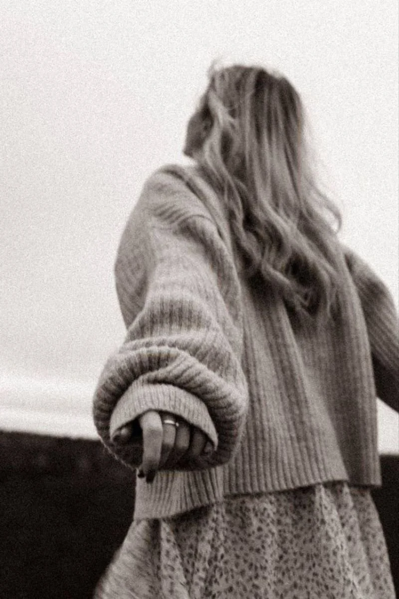 A woman with wavy hair wearing a chunky knit sweater and a patterned skirt, reaching out with her hand.