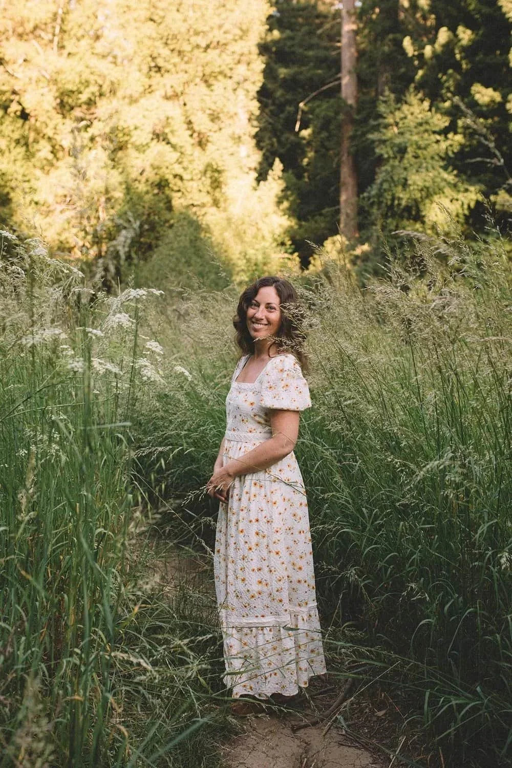 Female founder Nisa Brown in a white dress with yellow flowers stands on a dirt path surrounded by tall green grass, smiling in a forest with sunlight filtering through trees.