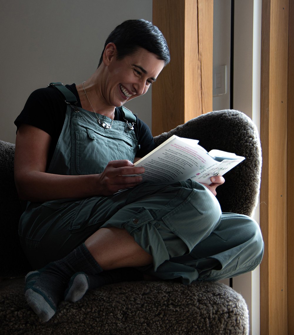 A person sitting on a couch, reading a book and smiling, while wearing a black shirt, green overalls, and gray socks near a wooden window frame.