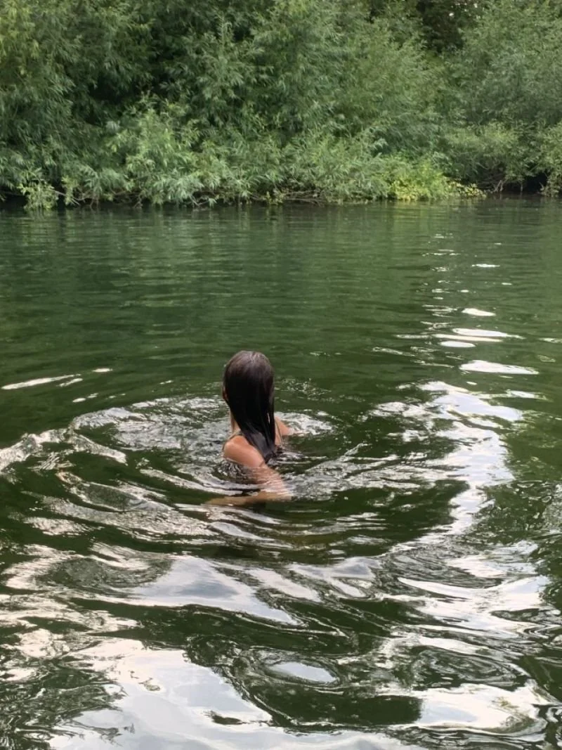 A person with long, dark hair swimming in a green, natural body of water, surrounded by lush trees and foliage.