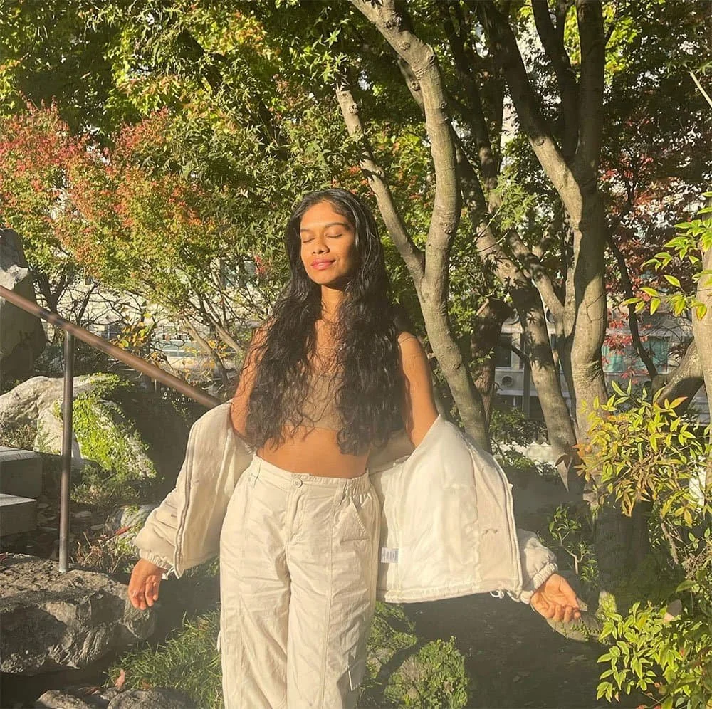 A woman with long dark curly hair standing outdoors in a garden with green and reddish foliage, wearing a beige crop top, beige pants, and a beige jacket draped off her shoulders, with eyes closed and a peaceful expression.