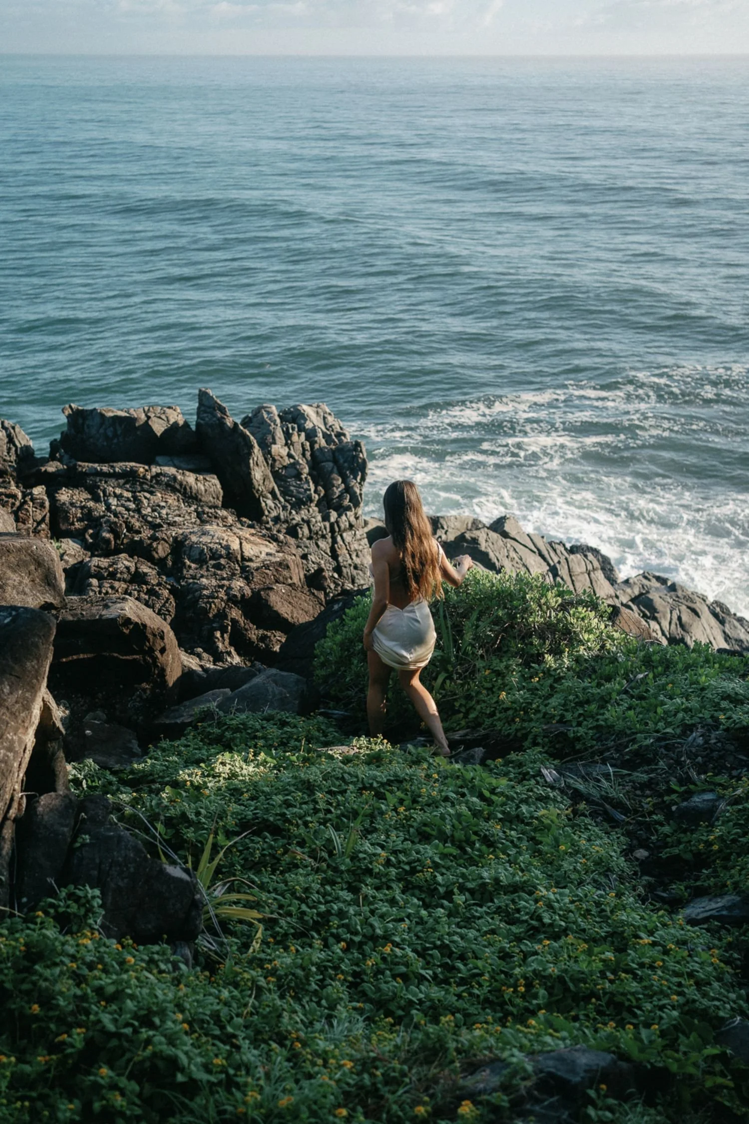 A woman with long brown hair wearing a white top and white shorts stands on rocky shore near green foliage, facing the ocean with waves and a distant horizon.