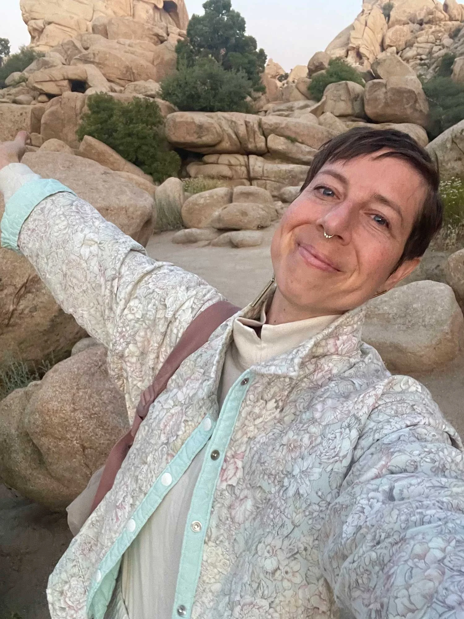 A smiling person with short dark hair, wearing a patterned jacket and a septum ring, taking a selfie in a rocky desert landscape with large boulders and sparse green vegetation.