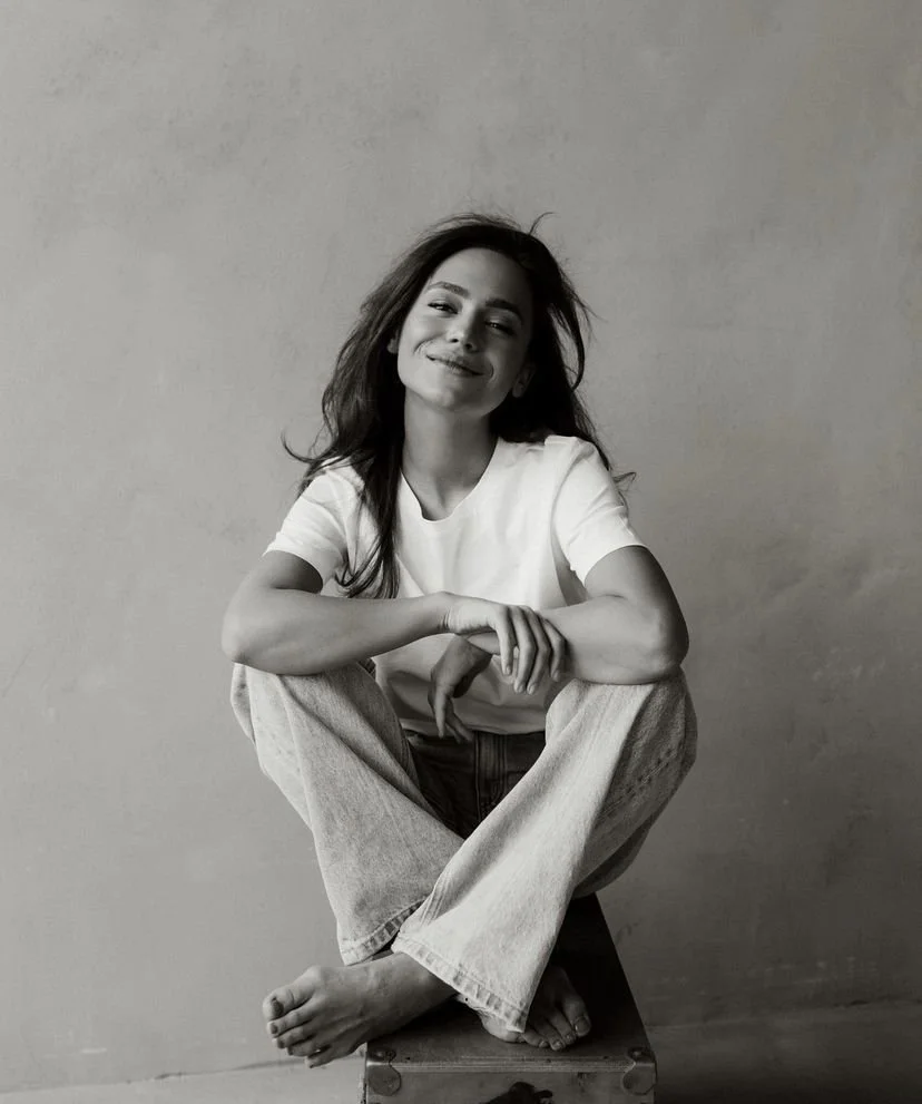 A woman sitting cross-legged on a small wooden stool, smiling softly at the camera, wearing a white t-shirt and light-colored jeans, with her hair loose and slightly tousled, against a plain wall.
