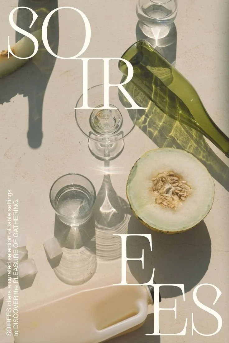 A picnic table setting with a halved melon, green leafy vegetable, clear glasses, and sunlight casting shadows.