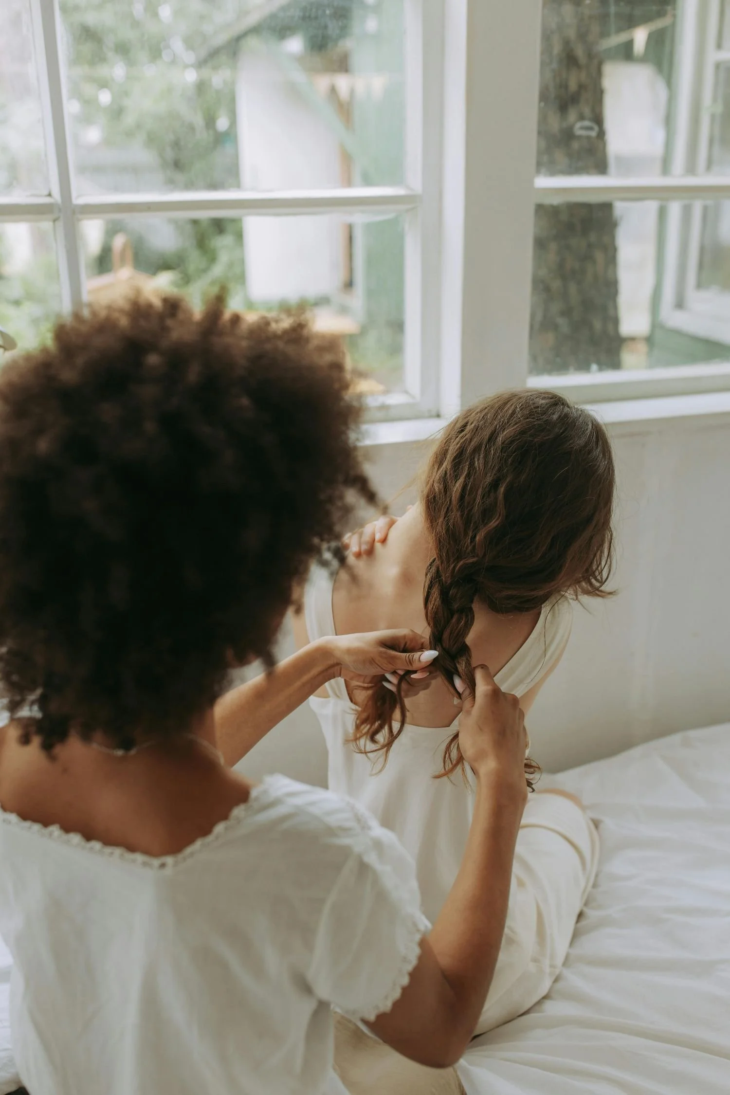 A woman is sitting on a bed by a window while another person is braiding her long, wavy brown hair.