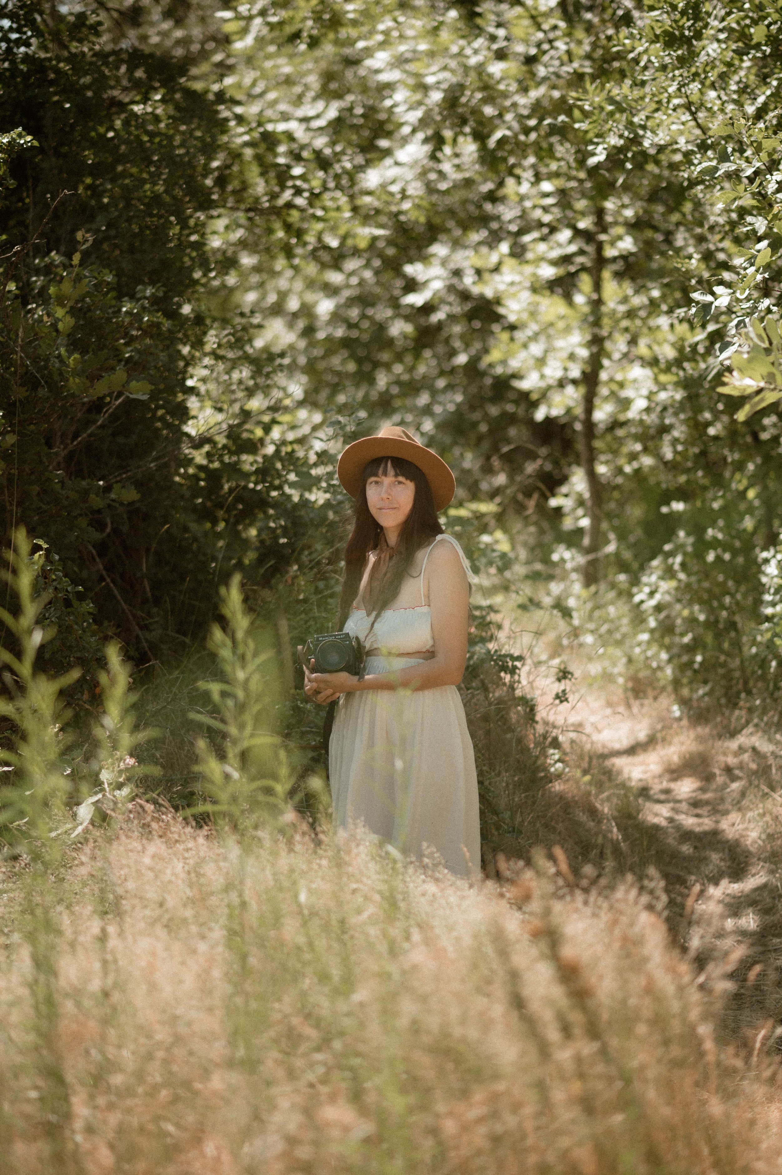 A woman with dark hair, wearing a wide-brimmed hat and a sleeveless dress, holding a vintage camera, standing in a wooded area with sunlight filtering through the trees.