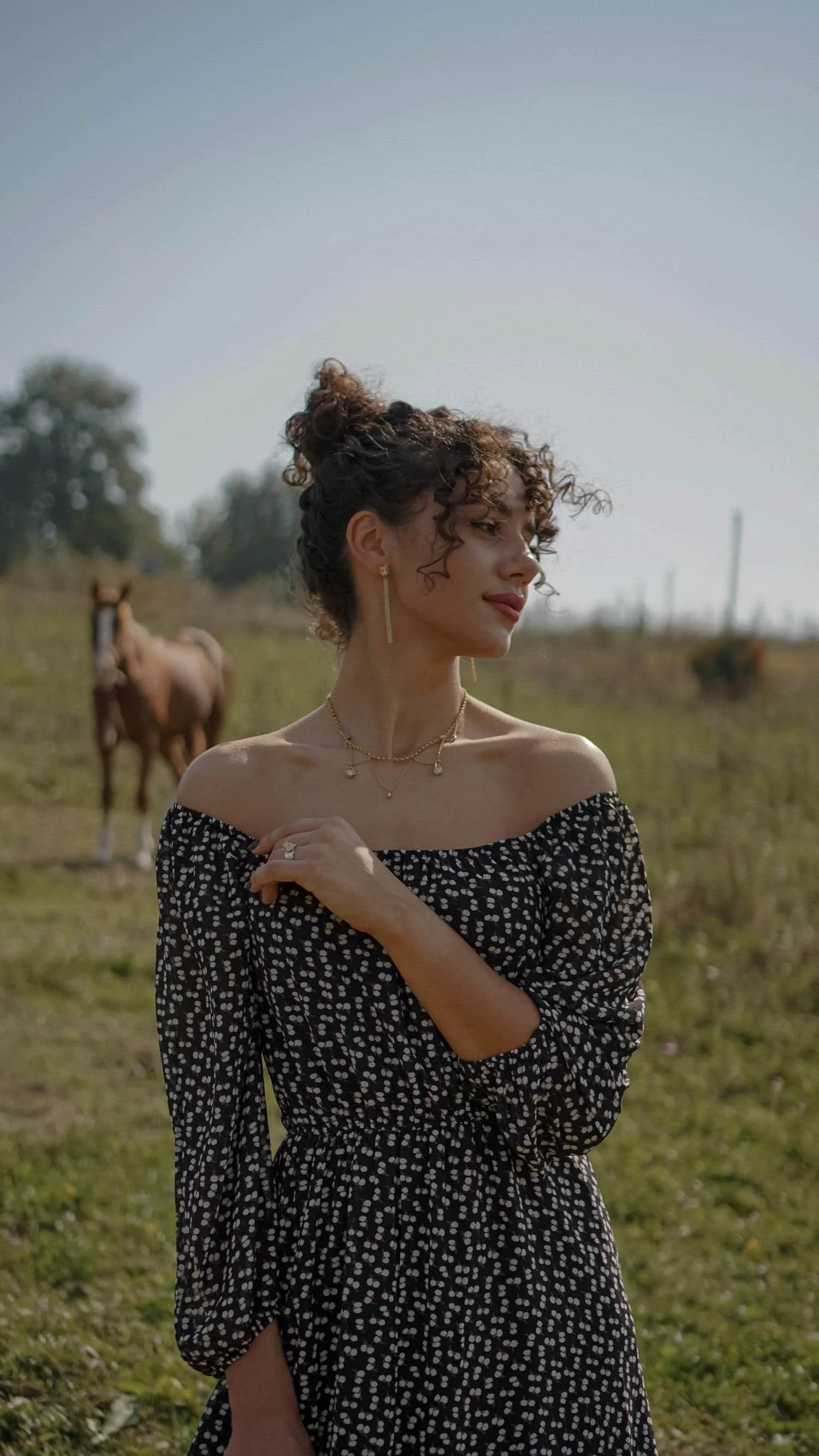A woman with curly hair in a bun wearing an off-shoulder black dress with white polka dots, layered gold necklaces and earrings, standing in a field with two horses in the background under a clear sky.