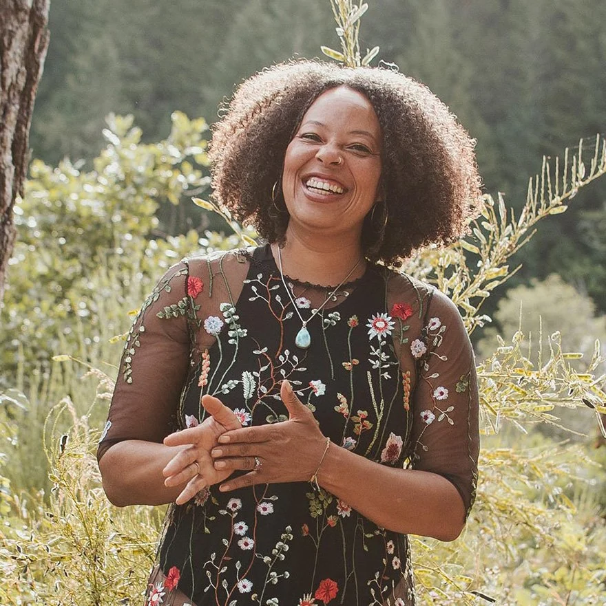 Smiling woman with curly hair wearing a floral embroidered sheer dress outdoors in a sunny, green, forested area.