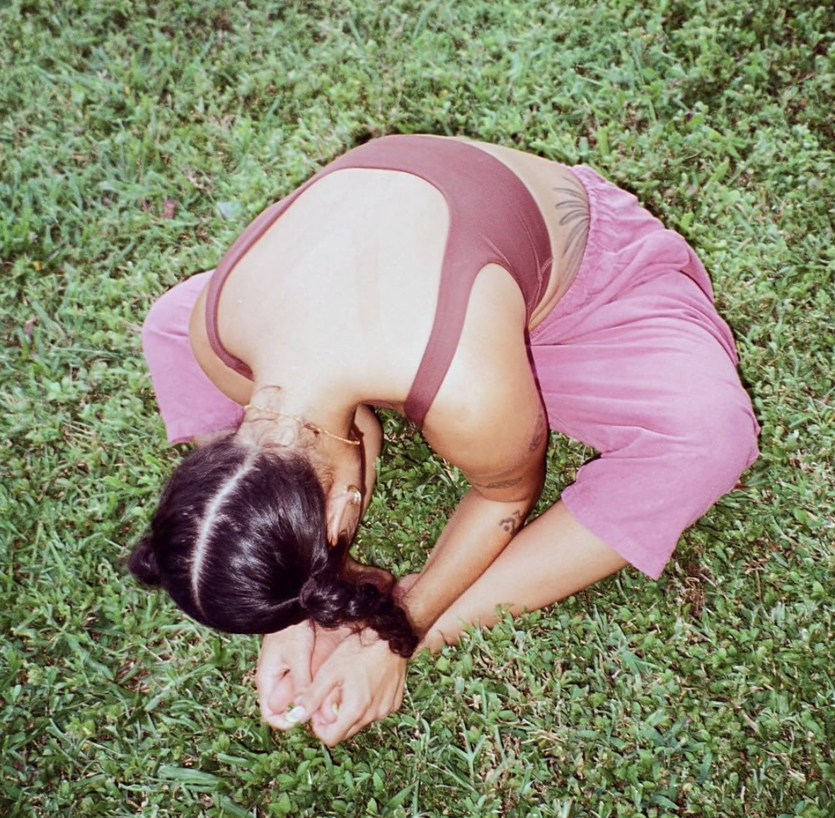 Person practicing yoga outdoors on green grass, wearing a tank top and pink pants, with hair braided and tattoos visible.