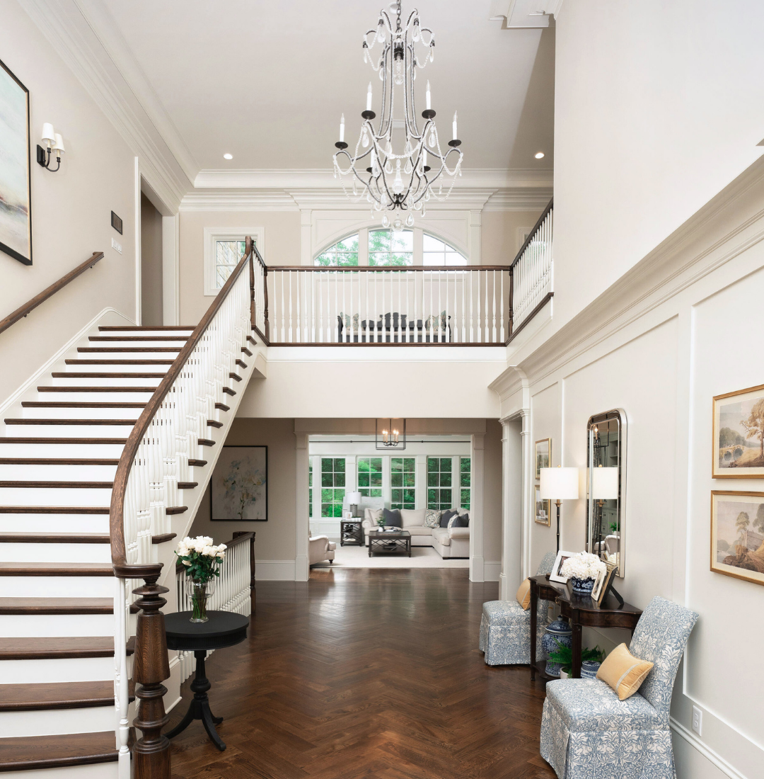 Spacious living room with staircase, chandelier, and large windows, decorated in neutral tones with elegant furniture and artwork.