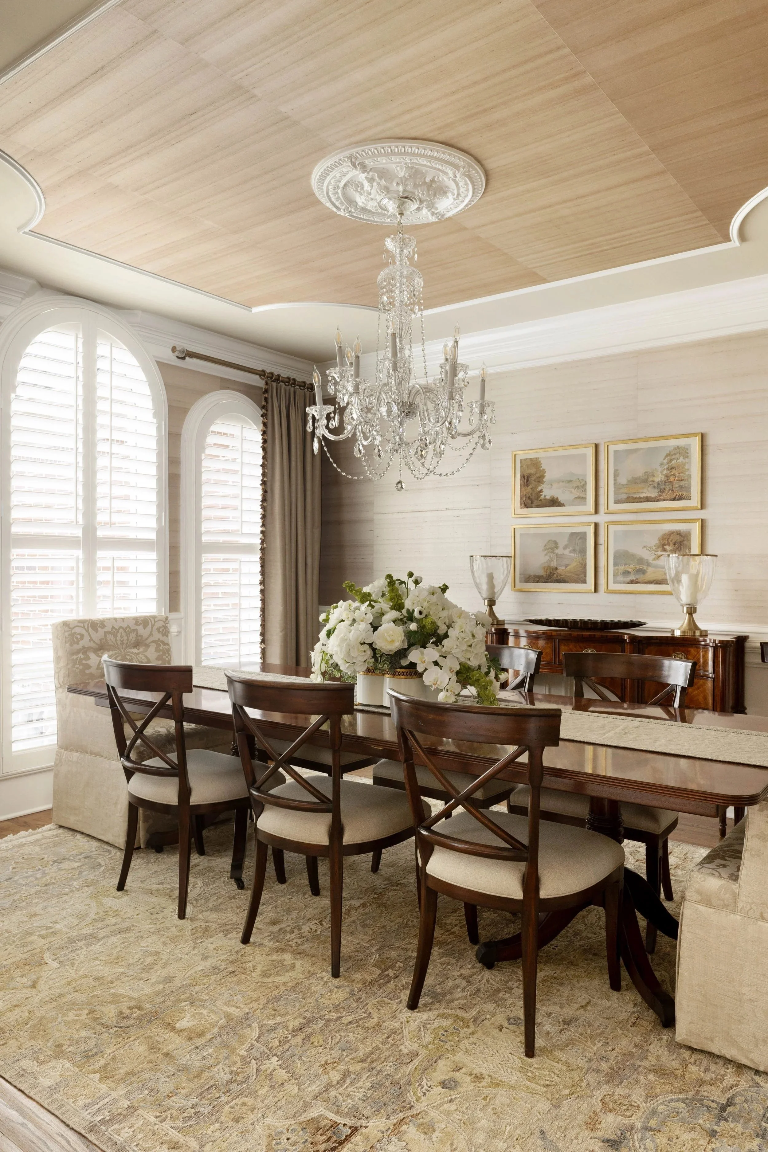 A dining room with a large wooden table surrounded by six dark wooden chairs with beige upholstered seats. On the table, there is a white floral centerpiece. The room features large, arched windows with white plantation shutters and draped curtains. A crystal chandelier hangs from the ceiling. The walls are decorated with four landscape paintings in gold frames, and there are two white lamps on a sideboard. The room has a beige patterned rug on the floor.