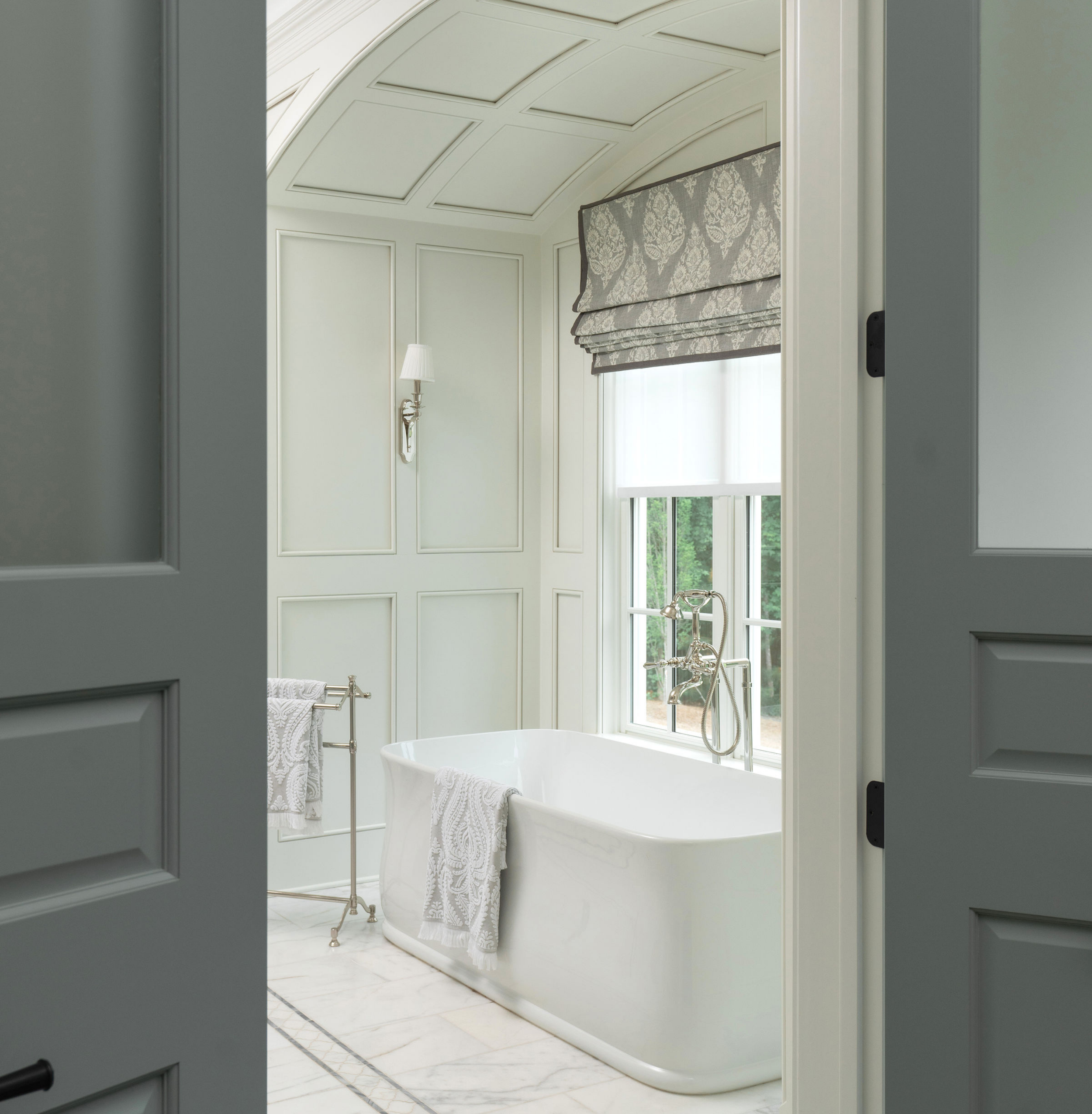 View from doorway into a bathroom with a freestanding bathtub next to a window with a patterned Roman shade, white-paneled walls, and a marble floor.