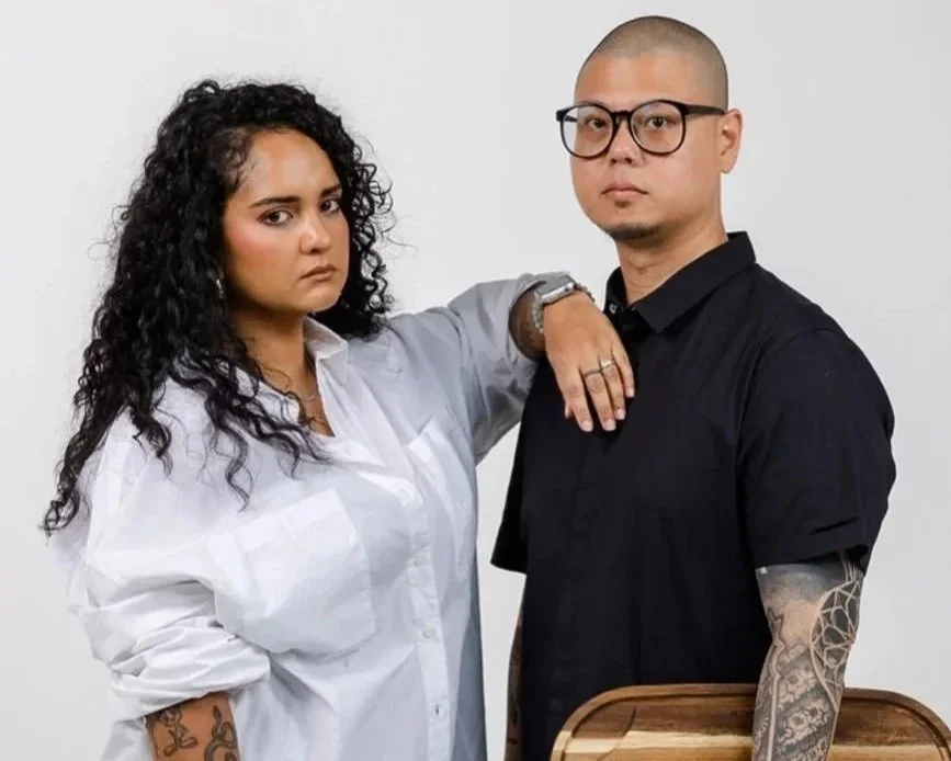 A woman with curly black hair and tattoos on her arm, wearing a white shirt, stands next to a bald man with glasses and tattoos, wearing a black shirt, against a white background.
