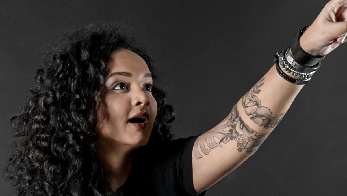 Woman with curly black hair, tattoos on her arm, and bracelets, raising her arm with a surprised expression against a dark background.