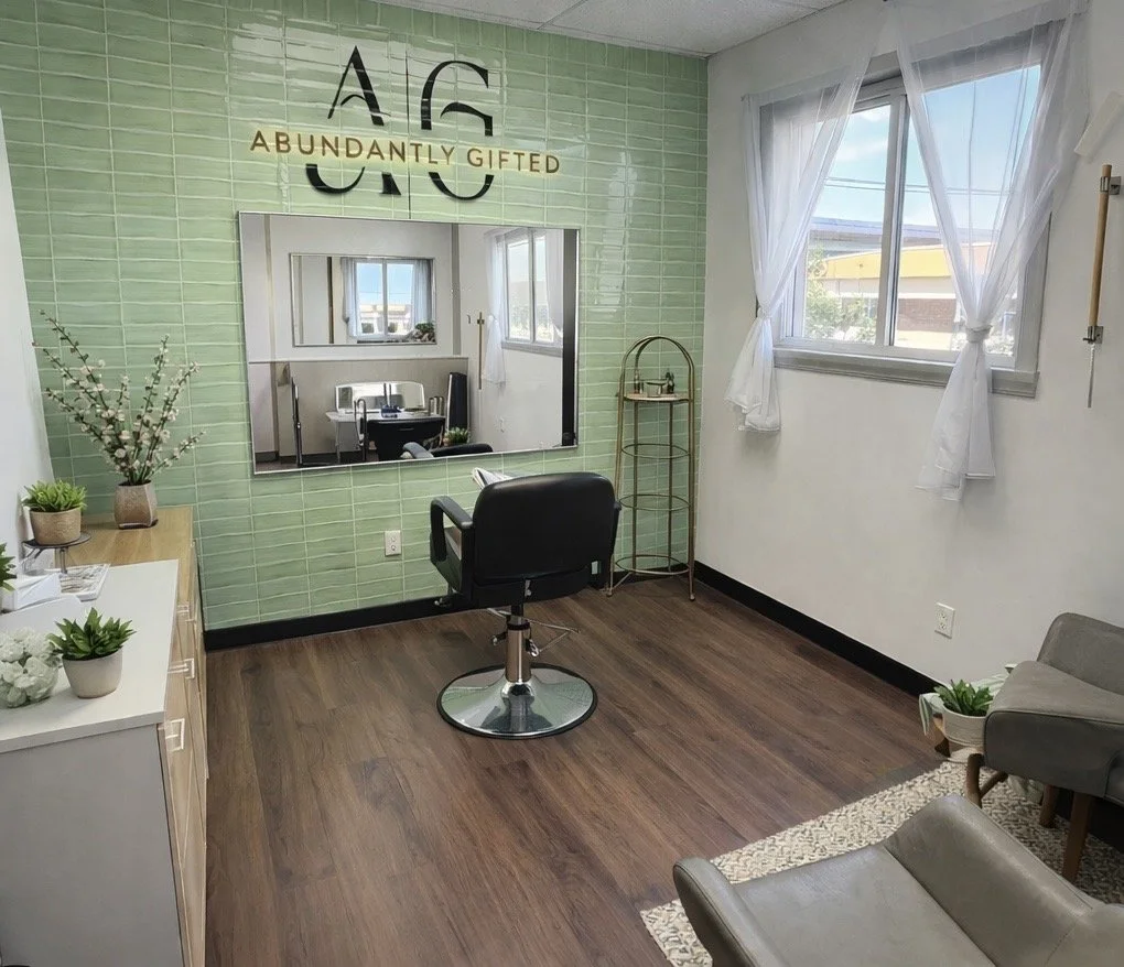 A salon or beauty studio with a large window, green tiled accent wall, and a mirror. There is a black salon chair, a small white cabinet with potted plants, and a beige armchair. The wall behind the mirror has an artistic sign that reads 'AG ABUNDANTLY GIFTED.' The room has wood flooring, sheer white curtains, and decorative plants.