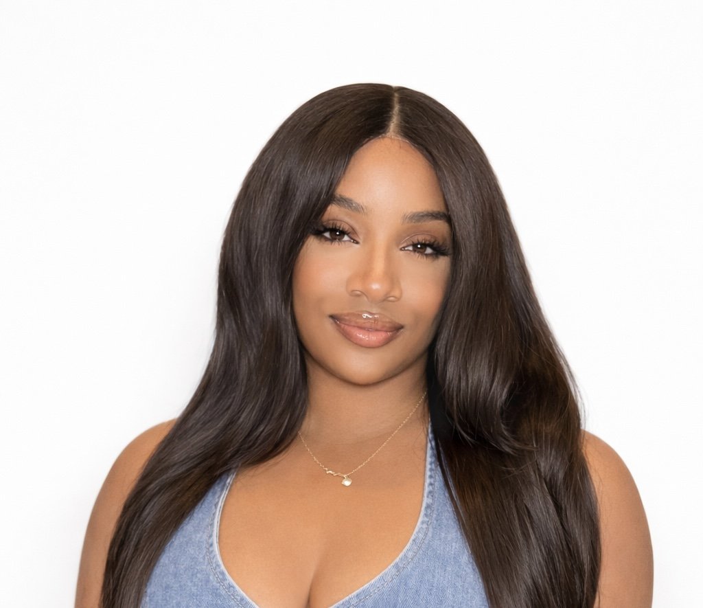 Portrait of a young woman with long dark hair, wearing a sleeveless denim top and a delicate necklace, smiling softly against a plain white background.